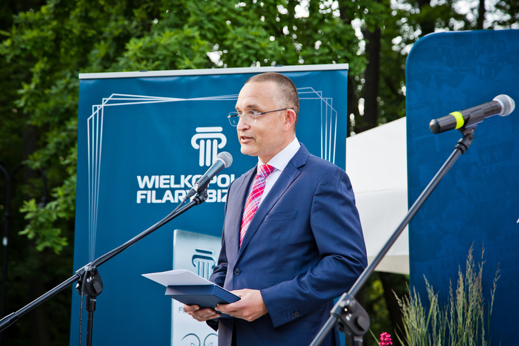 Mężczyzna w okularach, w granatowym garniturze i krawacie, przemawia do mikrofonu, trzymając ozdobne etui z dokumentem. W tle niebieska ścianka z napisem "Wielkopolskie Filary Biznesu".