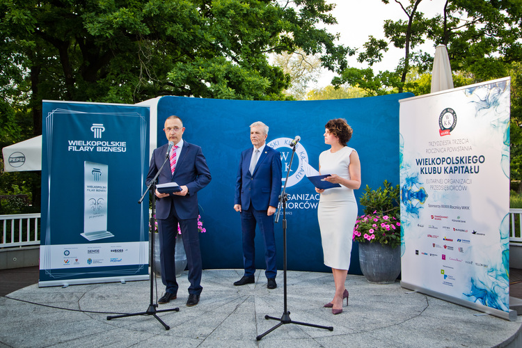 Trzy osoby w eleganckich strojach stoją na okrągłym, kamiennym podium. Mężczyzna po lewej przemawia do mikrofonu, obok niego stoi mężczyzna w niebieskim garniturze, a po prawej kobieta w białej sukience. W tle zielone drzewa, niebieska ścianka z logo oraz dwa roll-upy z informacjami Wielkopolskiego Klubu Kapitału.