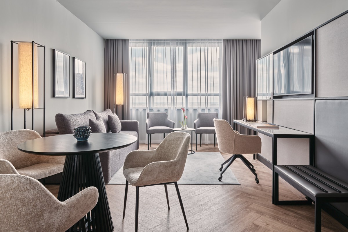 Nowoczesny apartament hotelowy z okrągłym stołem jadalnianym, szarą sofą i biurkiem, utrzymany w stonowanych odcieniach szarości, beżu i czerni z podłogą w jodełkę.