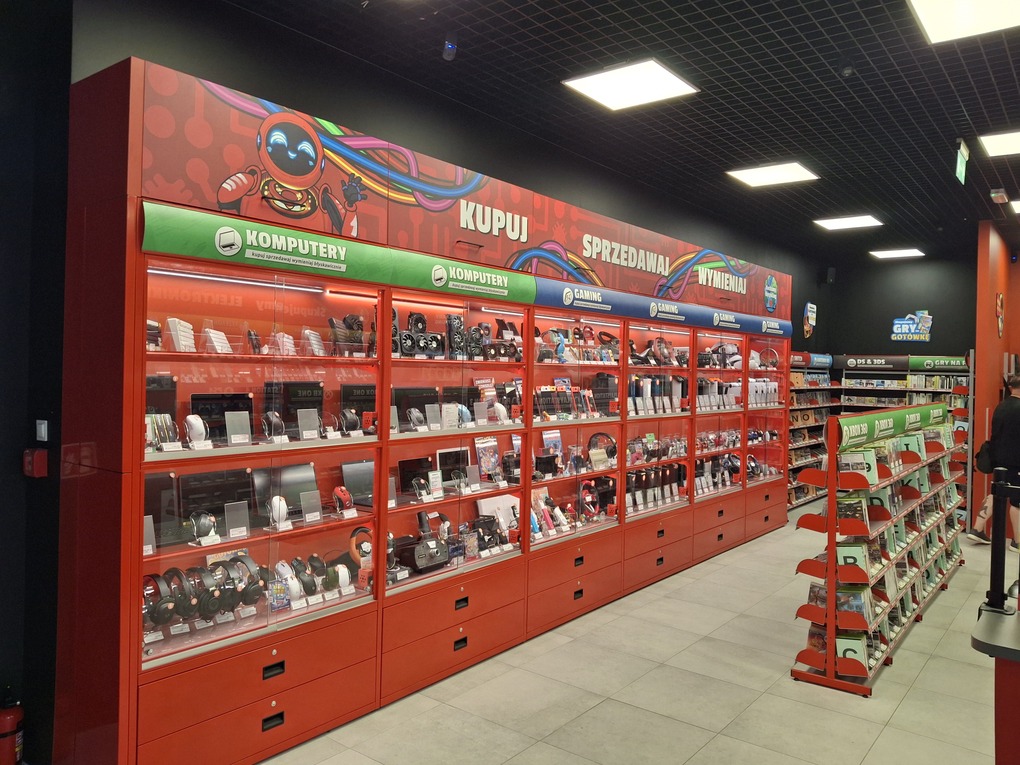 Wnętrze sklepu z elektroniką, dominują czerwone witryny pełne sprzętu komputerowego, nad nimi baner z napisem „KUPUJ SPRZEDAWAJ WYMIENIAJ”.