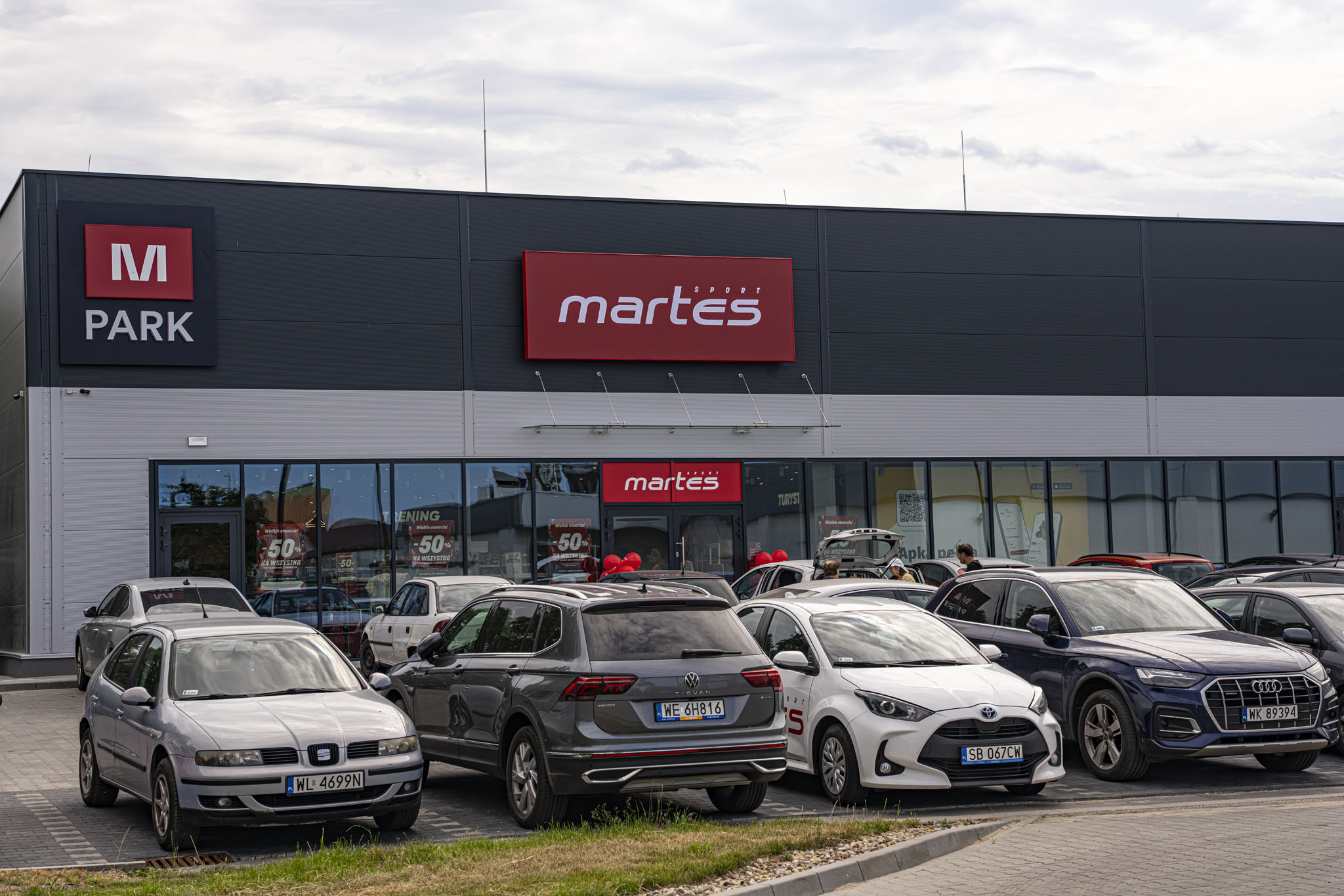 Nowoczesny budynek handlowy M Park z ciemną elewacją i czerwonym szyldem Martes Sport, przed którym znajduje się wypełniony parking.