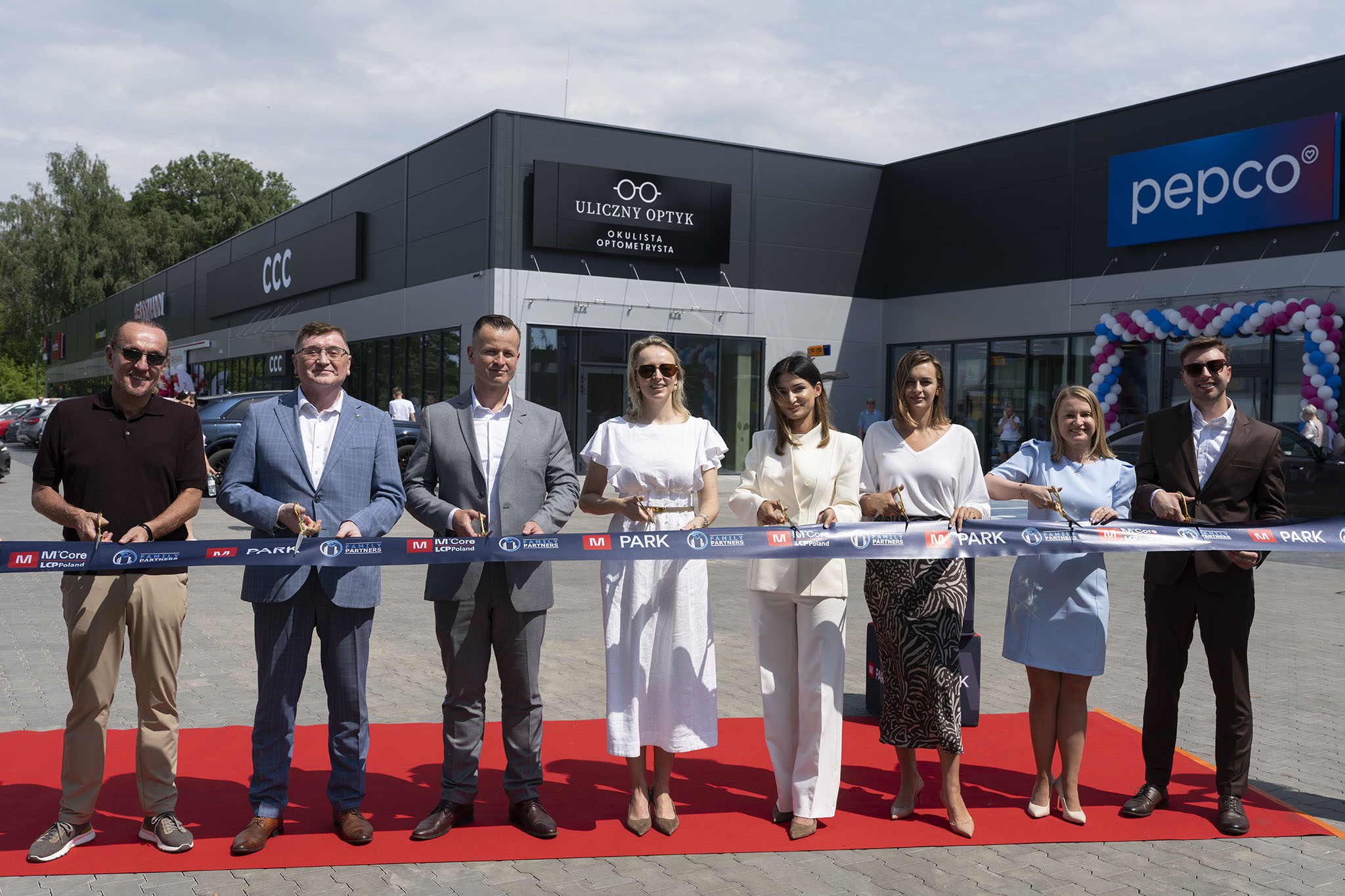 Grupa ośmiu elegancko ubranych osób przecina granatową wstęgę na czerwonym dywanie przed nowoczesnym pasażem handlowym M Park.