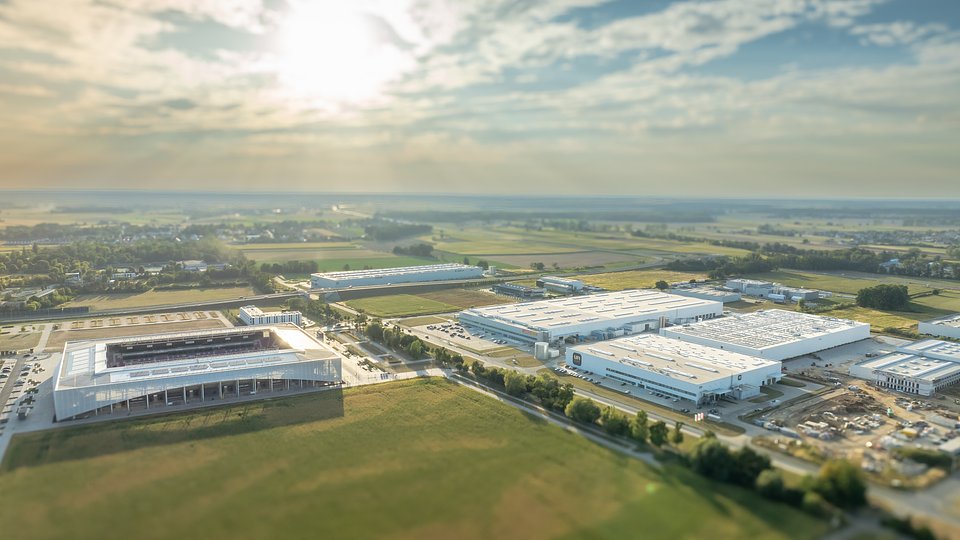 Widok z lotu ptaka na nowoczesny kompleks budynków przemysłowych, magazynów oraz stadion piłkarski na obrzeżach miasta.