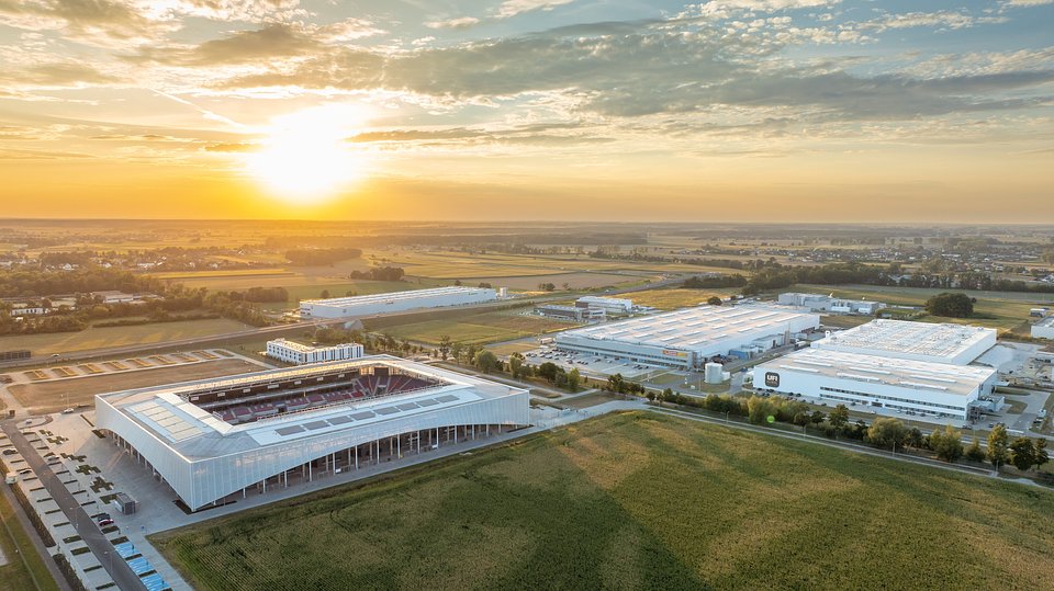 Widok z lotu ptaka przedstawia nowoczesny stadion piłkarski o białej elewacji, obok kompleks hal przemysłowych i logistycznych.
