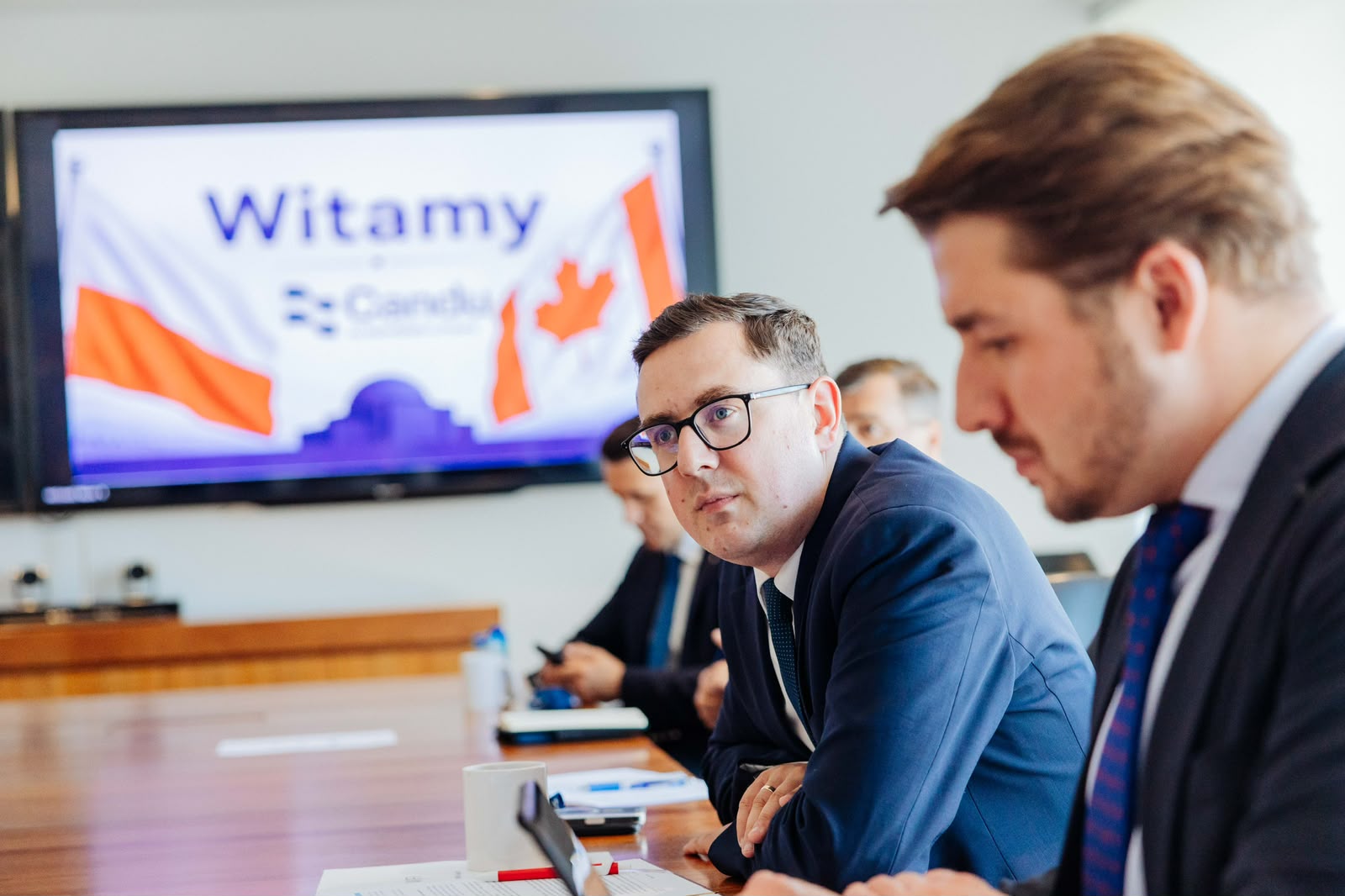 Mężczyźni w garniturach siedzą przy długim, drewnianym stole konferencyjnym, a w tle widać monitor z napisem „Witamy”.