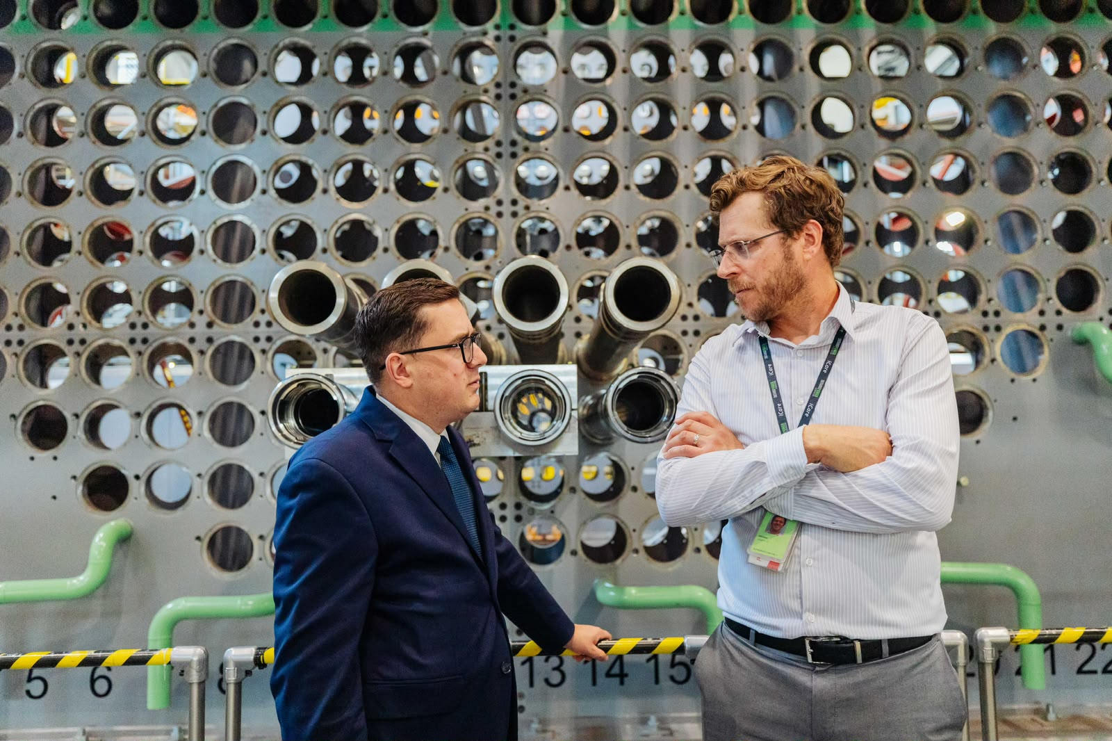 Dwaj mężczyźni prowadzą rozmowę na tle ogromnej, perforowanej metalowej ściany będącej częścią instalacji przemysłowej.