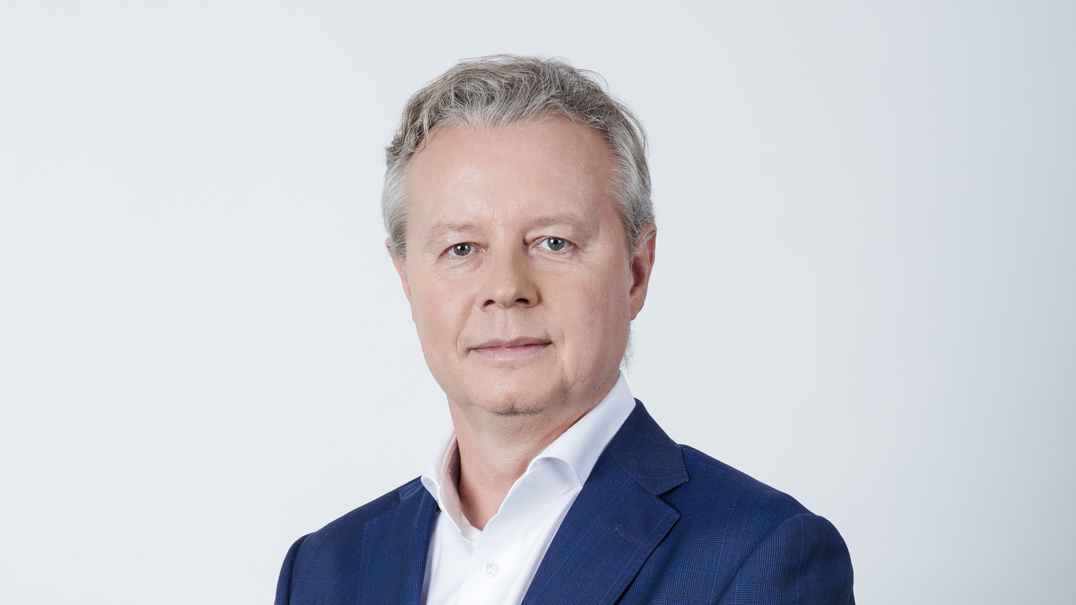 Portret mężczyzny o jasnych oczach i krótkich, siwych włosach, w białej koszuli i granatowej marynarce na szarym tle.