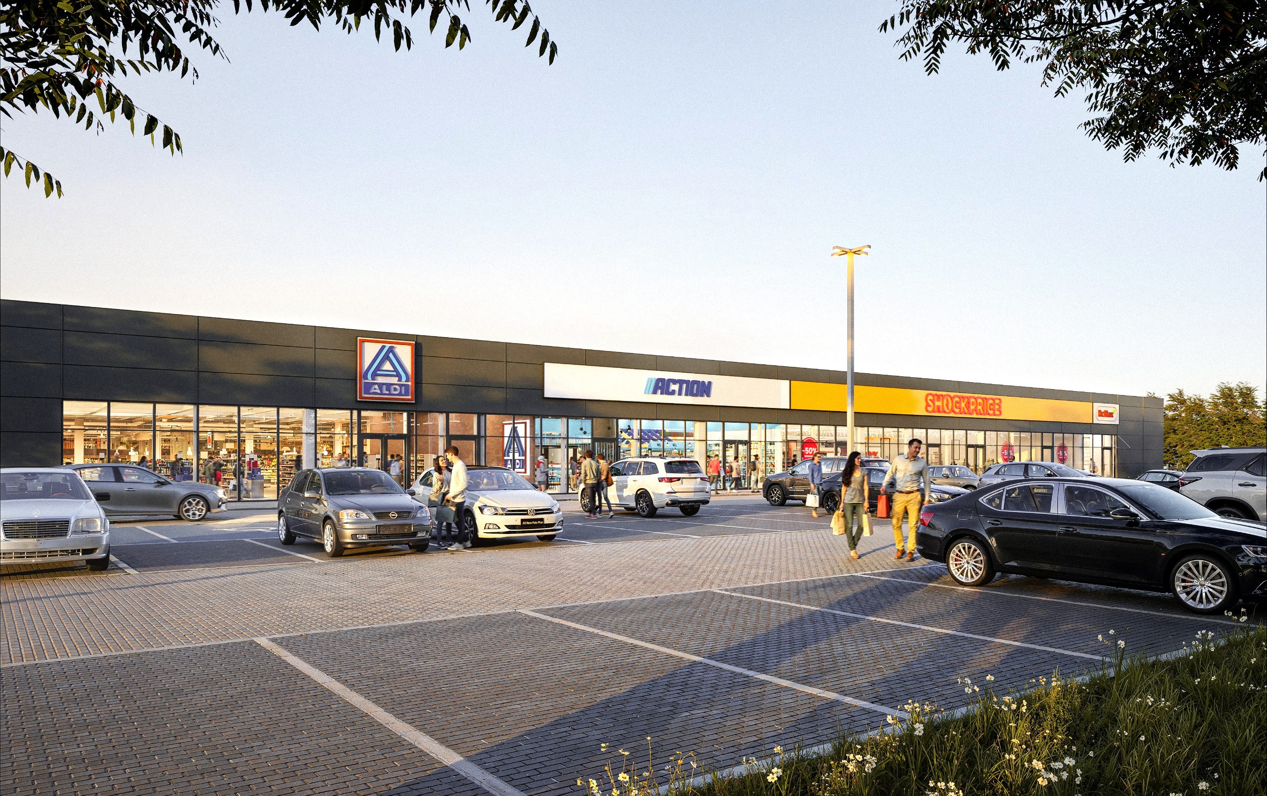 Nowoczesny park handlowy z ciemnoszarą elewacją i dużymi przeszkleniami. Przed budynkiem z logotypami Aldi, Action i Shockprice znajduje się rozległy parking z samochodami i pieszymi.