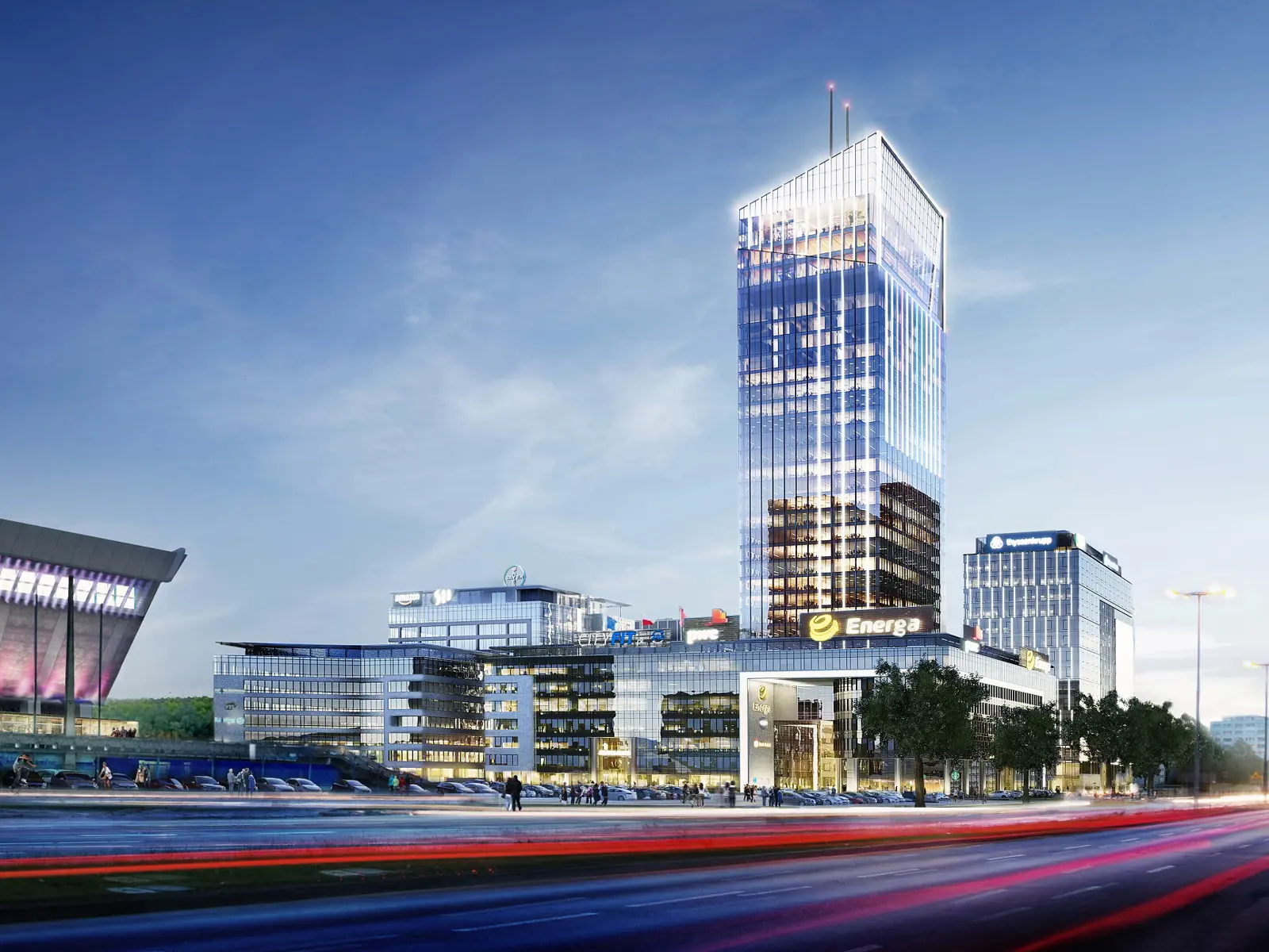 Nowoczesny kompleks biurowy z przeszklonym wieżowcem Energa, z elewacjami ze szkła i stali w niebieskich odcieniach, z widocznymi logo Amazon, Bayer, CityFit. Na pierwszym planie ulica z rozmytymi światłami samochodów o zmierzchu. miejska infrastruktura z drzewami przy chodnikach.