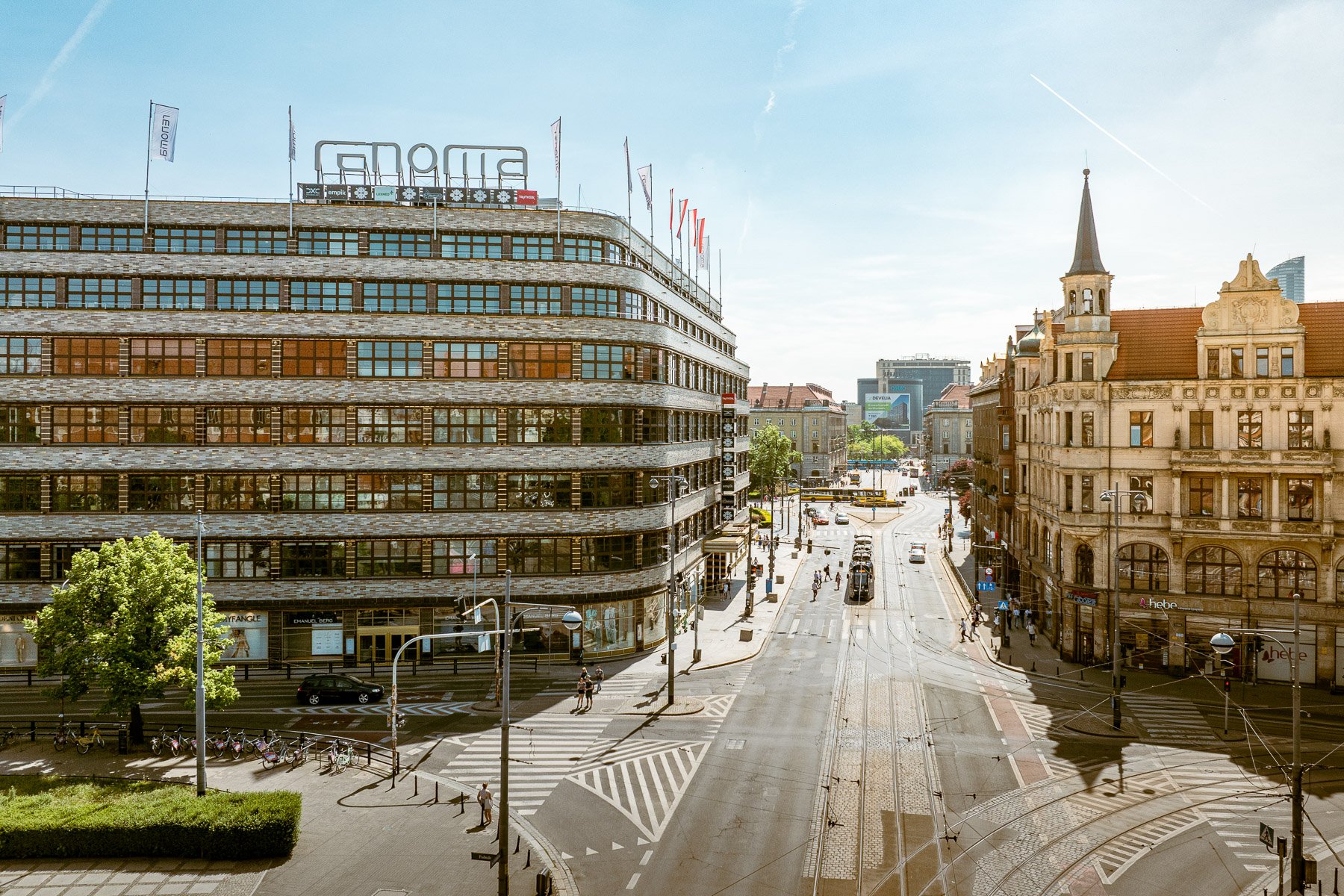 Wielkomiejska ulica z torowiskiem tramwajowym, otoczona modernistycznym domem towarowym Renoma i zabytkową kamienicą, w pełnym słońcu.