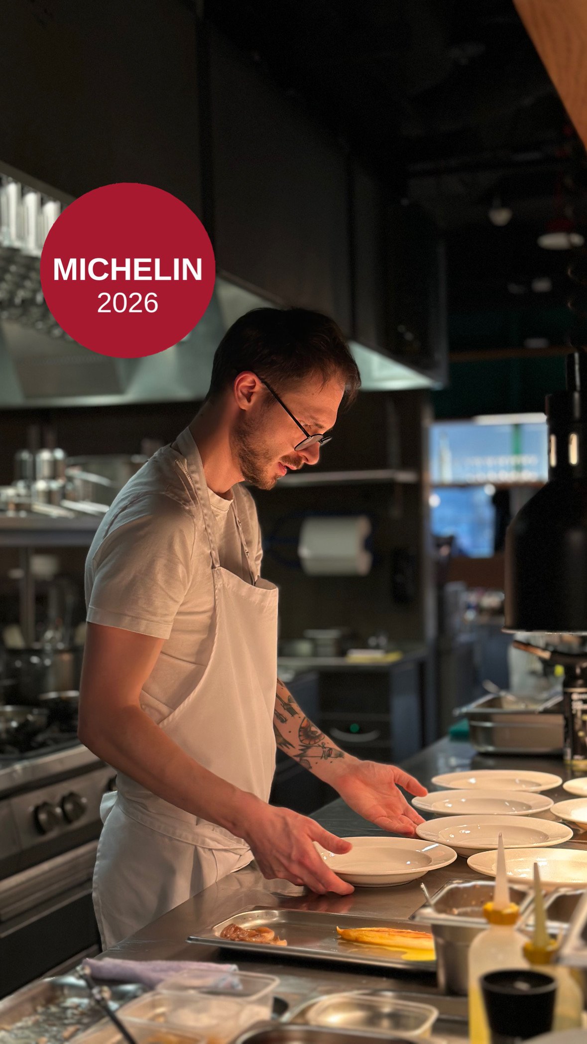 Mężczyzna w okularach i białym fartuchu kuchennym przygotowuje białe talerze na metalowym blacie w profesjonalnej kuchni, z czerwonym logo Michelin w tle.