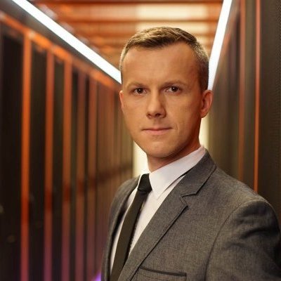 Portret mężczyzny o krótkich, ciemnych blond włosach, w szarej marynarce, białej koszuli, ciemnym krawacie, w nowoczesnym wnętrzu serwerowni z rzędami szaf rackowych.