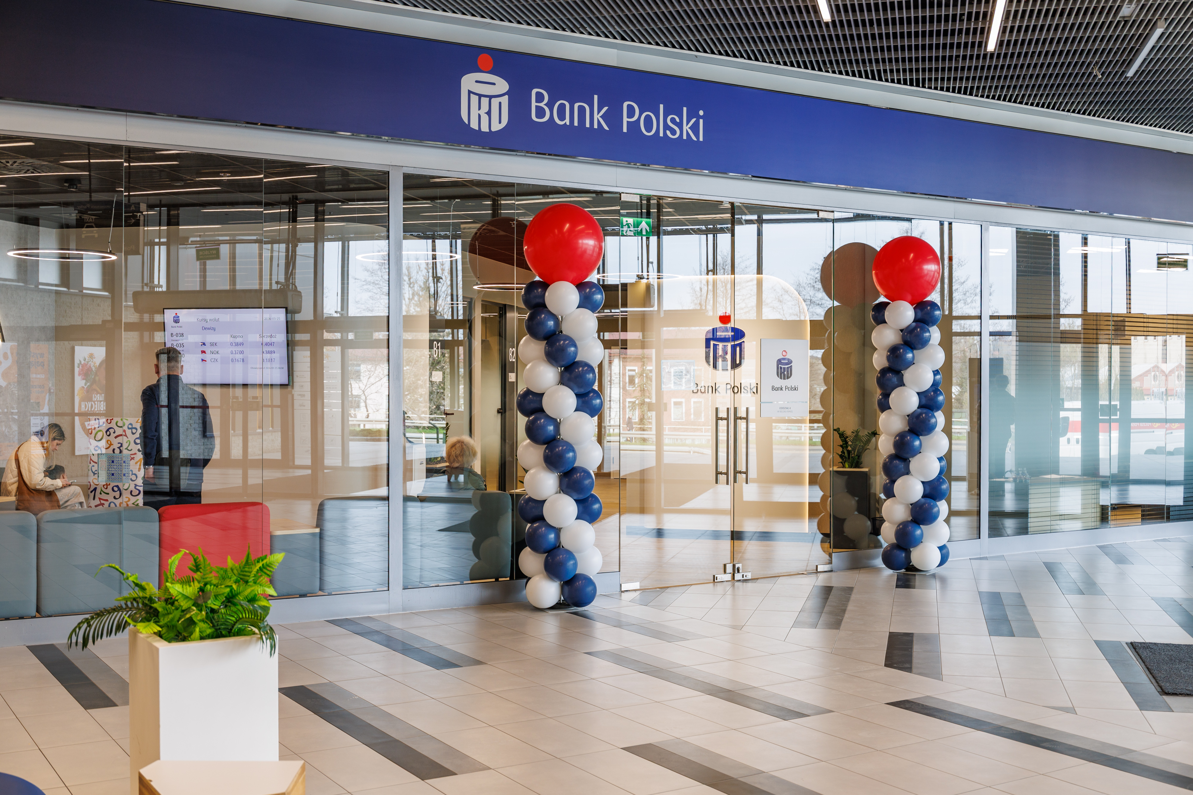 Nowoczesne i przeszklone wejście do placówki Banku Polskiego z granatowym szyldem i balonami. Wewnątrz klienci i ekrany.