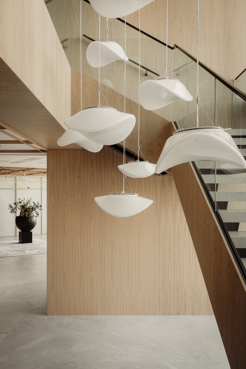 Nowoczesne wnętrze z klatką schodową ze szklaną balustradą i betonowymi stopniami. Dominują tu jasne drewno i kaskada zawieszonych, organicznych lamp.