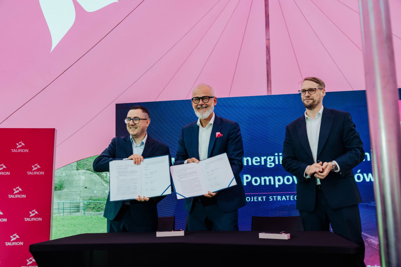 Trzej mężczyźni w garniturach uśmiechają się, stojąc za stołem pod namiotem, jeden z nich prezentuje dokumenty, widoczne logotypy Tauron.