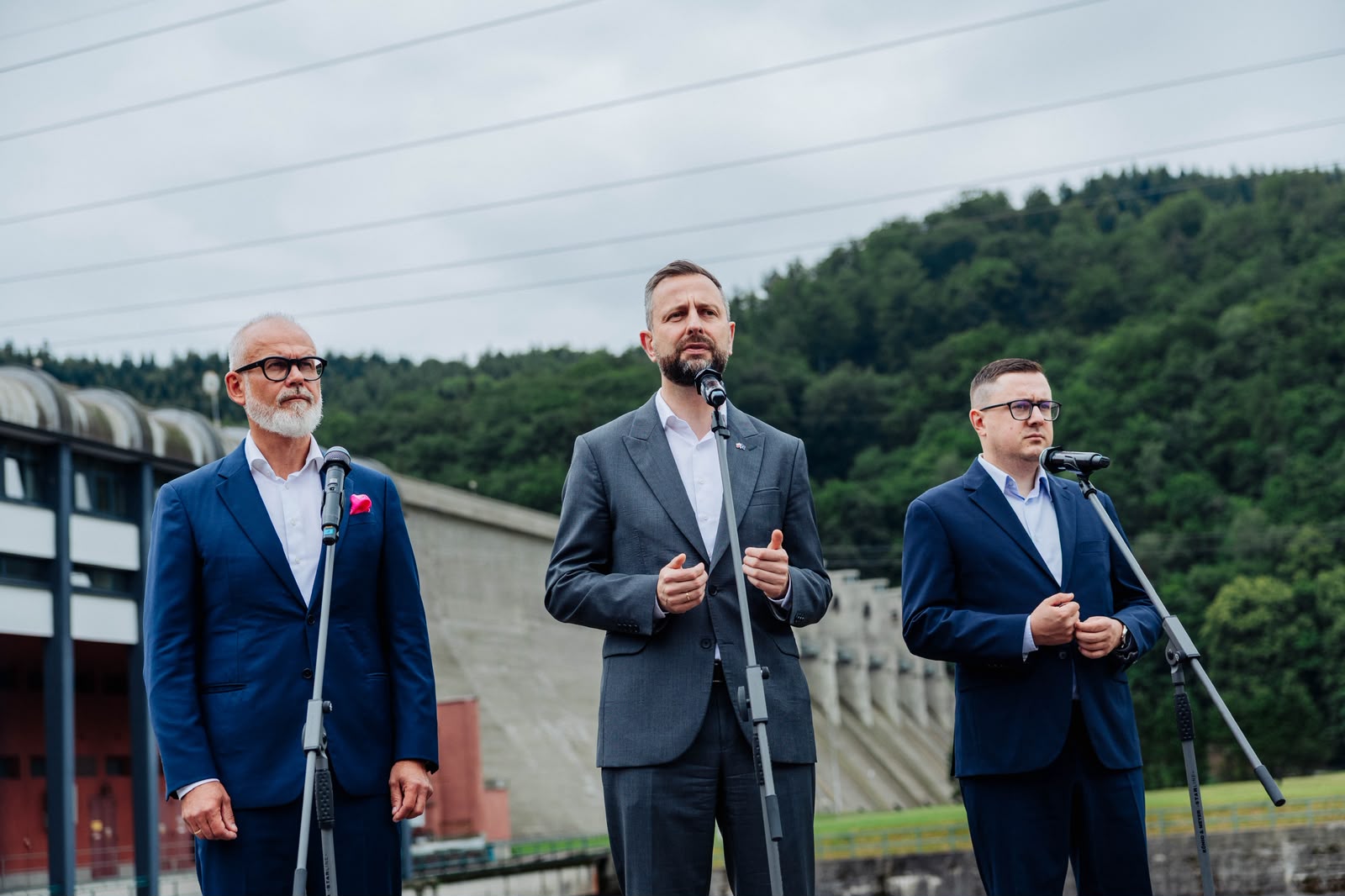 Trzej mężczyźni w garniturach stoją za mikrofonami, jeden przemawia, reszta słucha, na tle betonowej zapory wodnej zalesionego wzgórza.