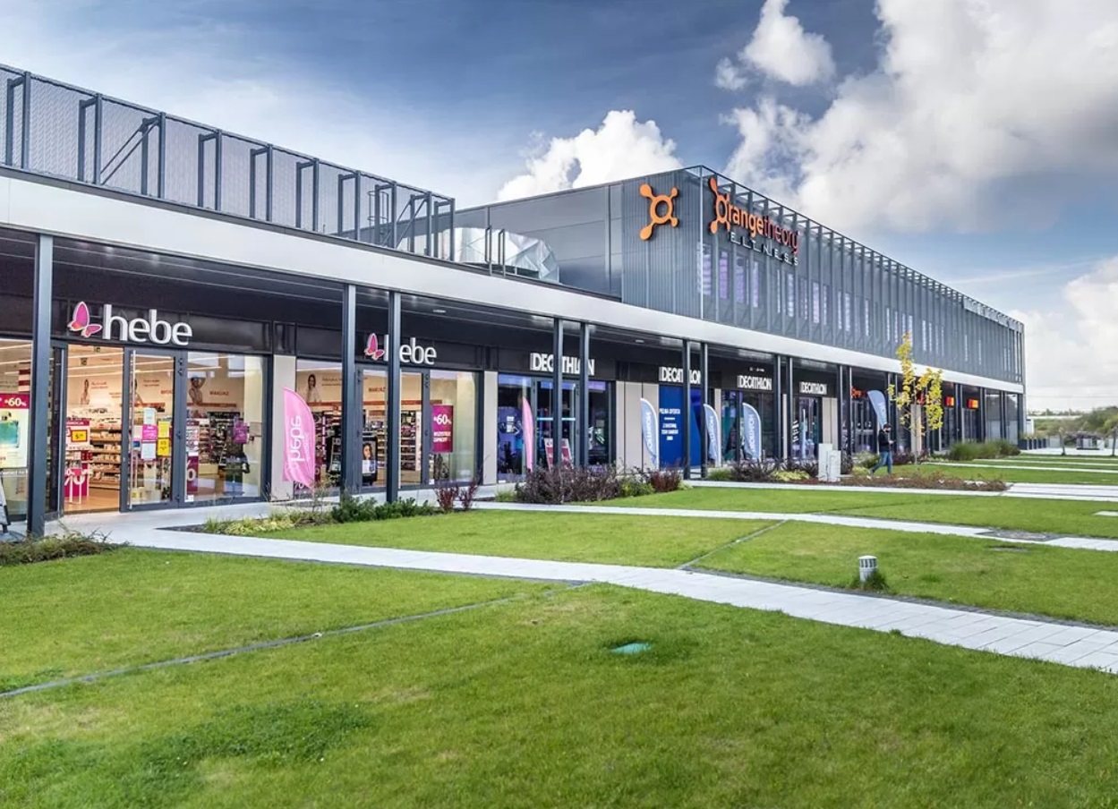 Nowoczesny, parterowy budynek handlowo-usługowy z ciemną elewacją i dużymi przeszkleniami, mieszczący Hebe, Decathlon i Orangetheory Fitness.