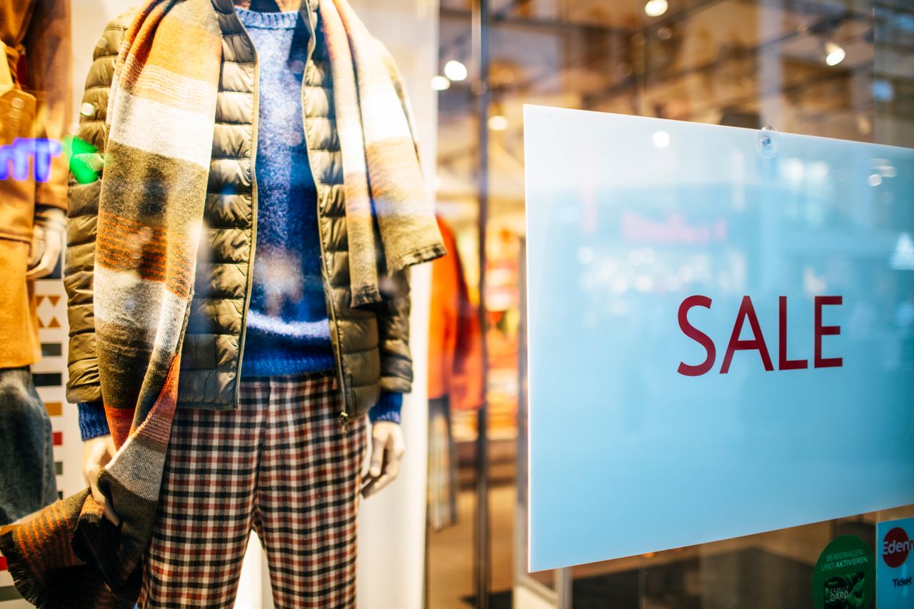 Manekin ubrany w pikowaną kurtkę, sweter, spodnie w kratę i pasiasty szal, obok widoczna tabliczka z napisem „SALE”.