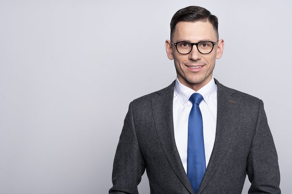 Uśmiechnięty mężczyzna w okularach o czarnych oprawkach, ubrany w ciemnoszary garnitur, białą koszulę i niebieski krawat, na jasnoszarym tle.