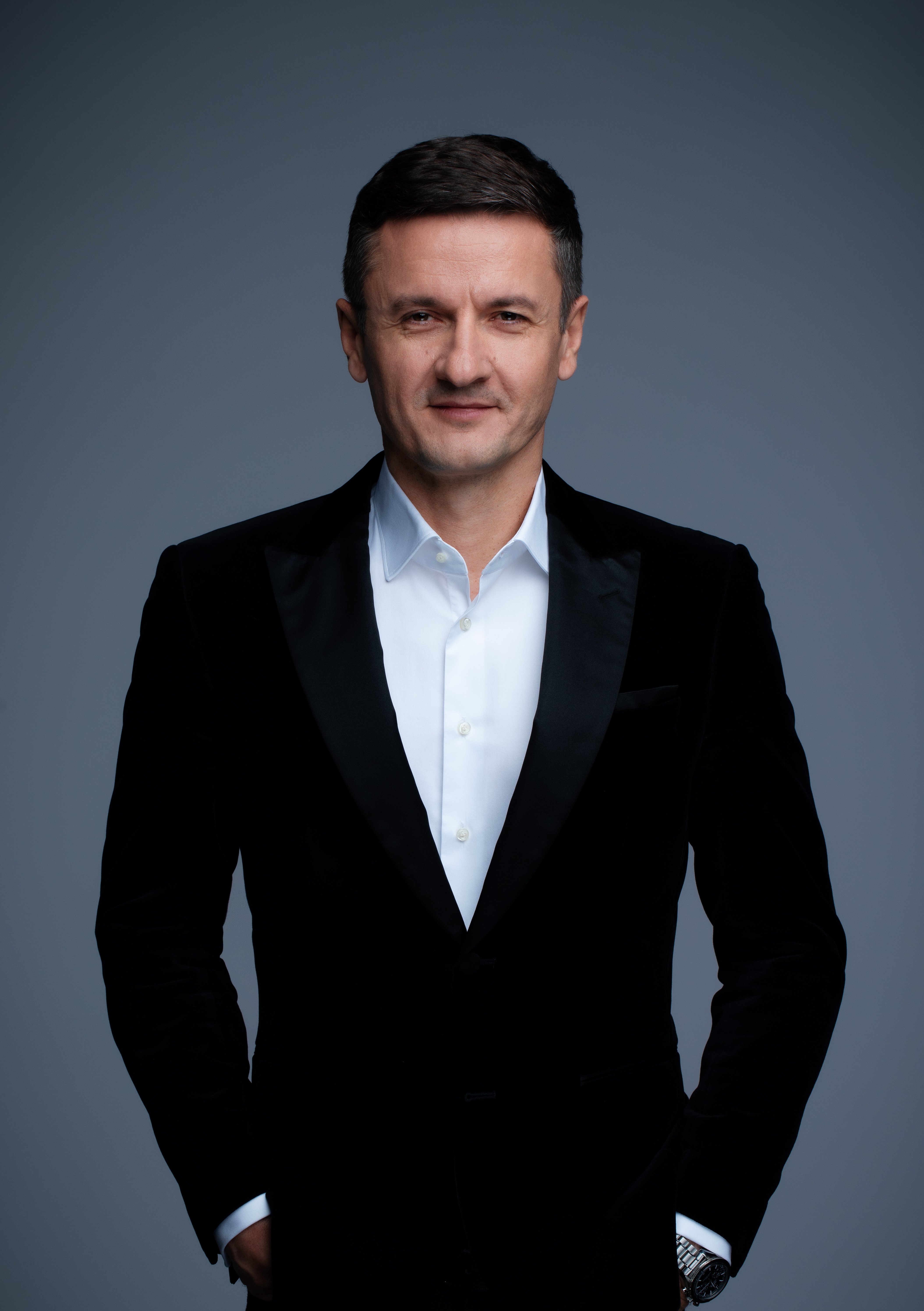 Mężczyzna w średnim wieku, o ciemnych, krótkich włosach, w białej koszuli i czarnej marynarce, patrzy w obiektyw.