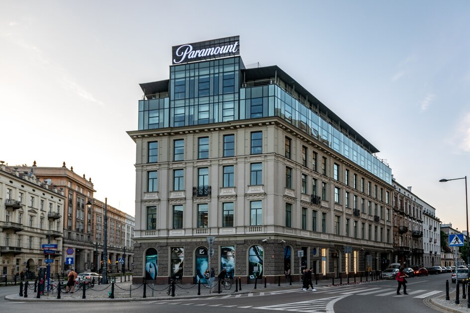 Narożna kamienica o jasnej elewacji łączy klasyczną architekturę z nowoczesną, przeszkloną nadbudową, na której widnieje logotyp „Paramount”.