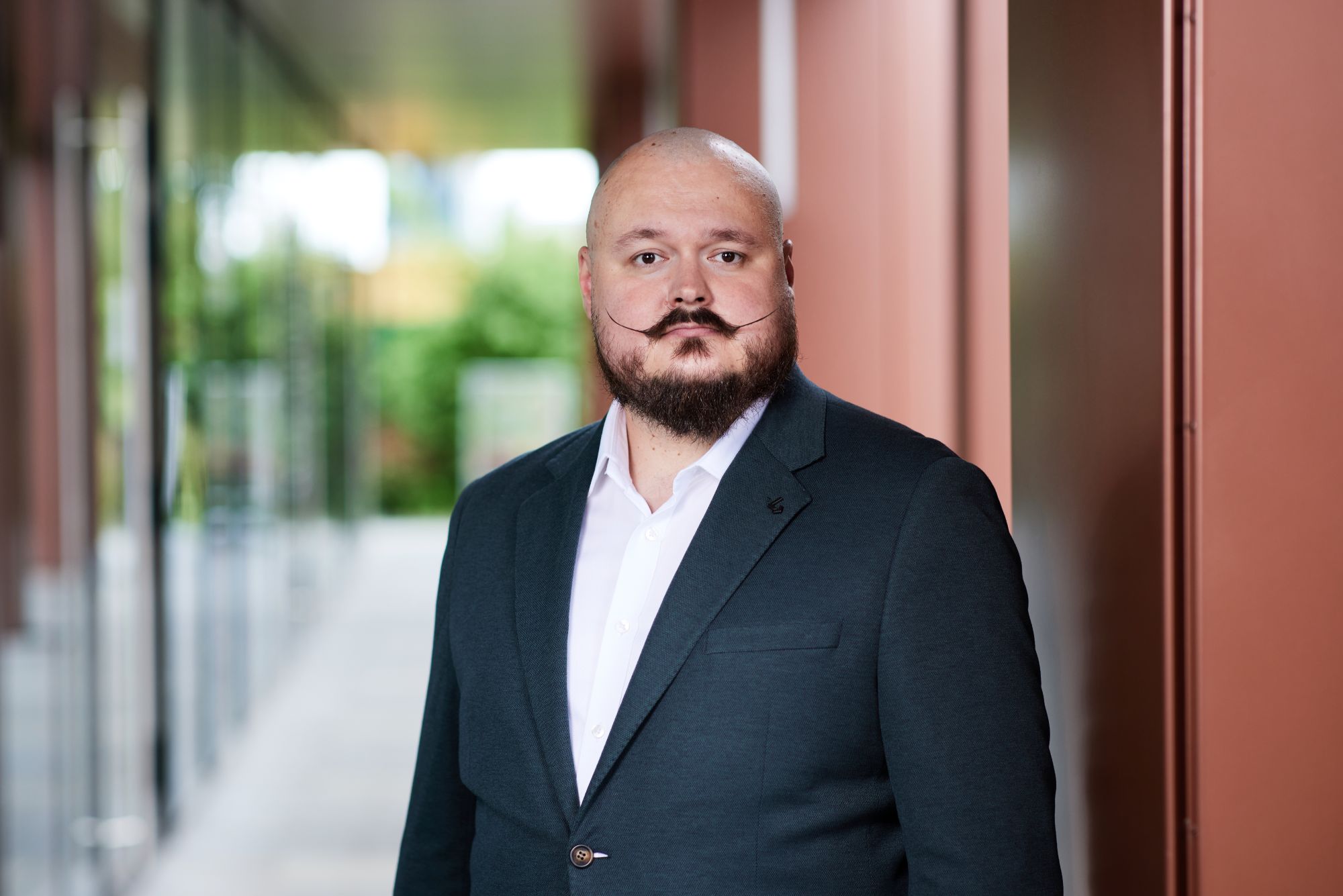 Portret uśmiechniętego mężczyzny z ogoloną głową, ciemną brodą i charakterystycznie podkręconymi wąsami, w ciemnej marynarce.