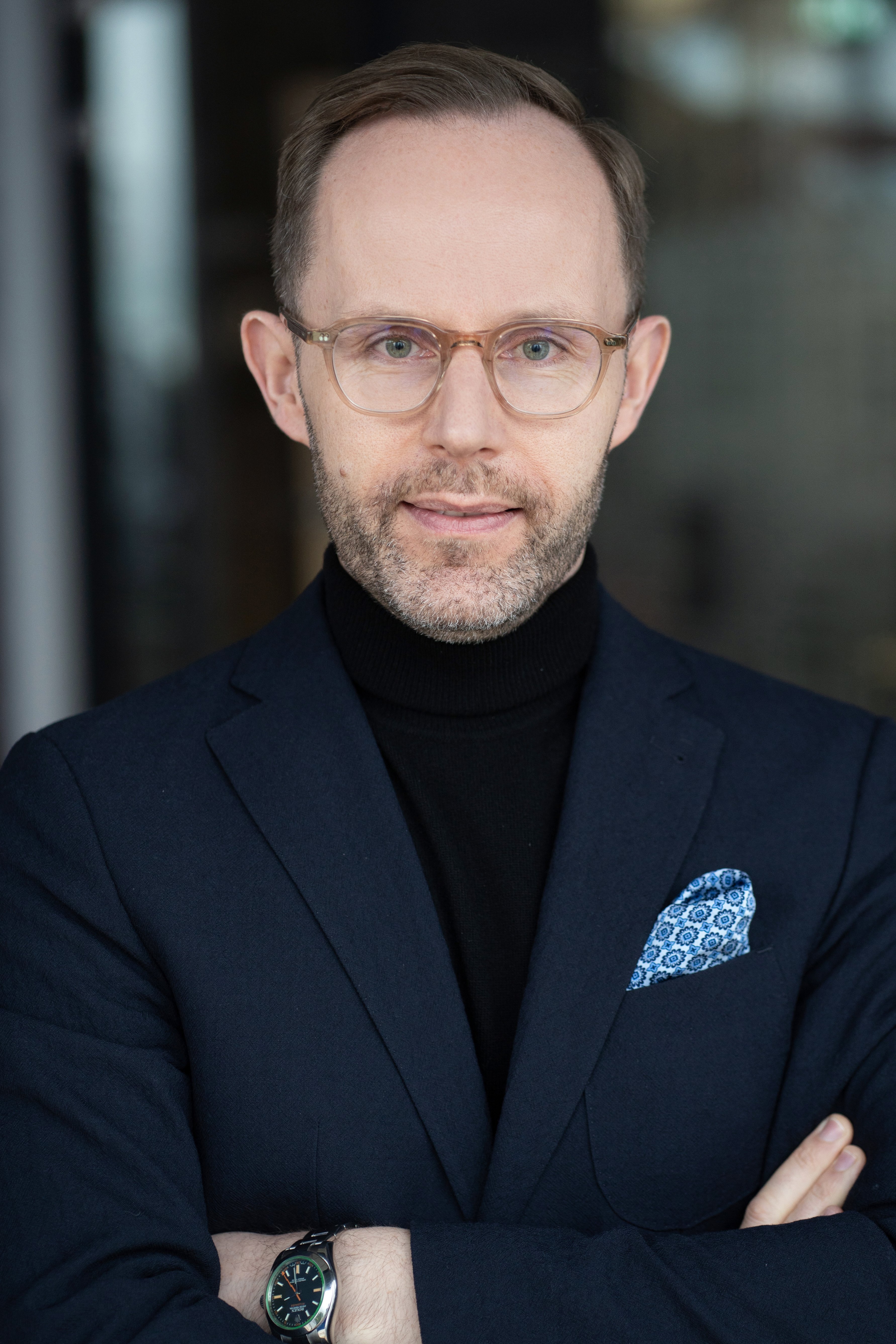 Portret mężczyzny w średnim wieku z krótkim zarostem i okularami, ubranego w granatową marynarkę, niebieską poszetkę i czarny golf. Postać stoi z założonymi rękami na rozmytym tle.