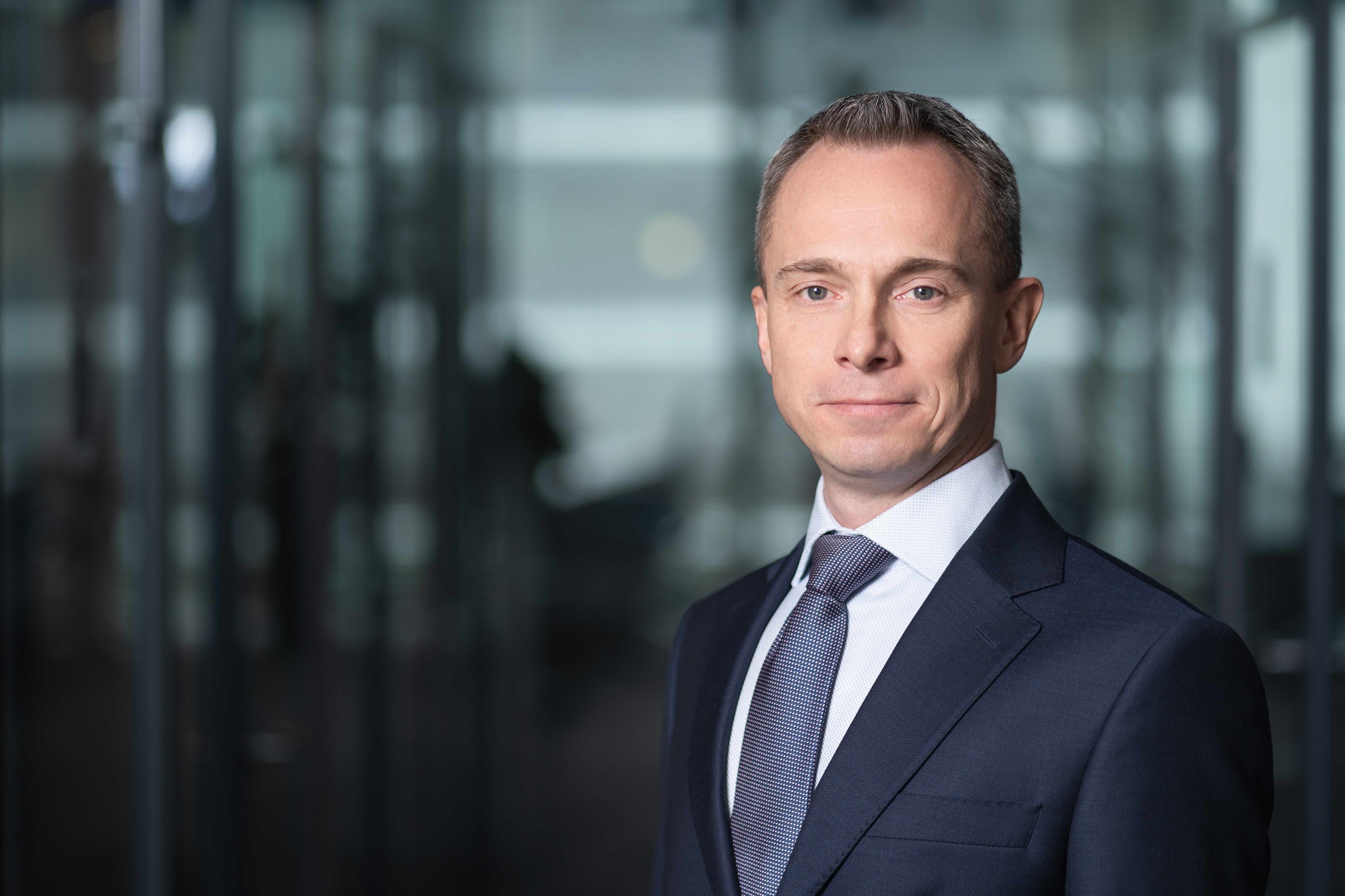 Portret mężczyzny w średnim wieku z krótkimi, szpakowatymi włosami, ubranego w ciemnogranatowy garnitur, jasną koszulę i wzorzysty krawat. Patrzy w obiektyw z lekkim uśmiechem na tle rozmytego biurowego wnętrza.