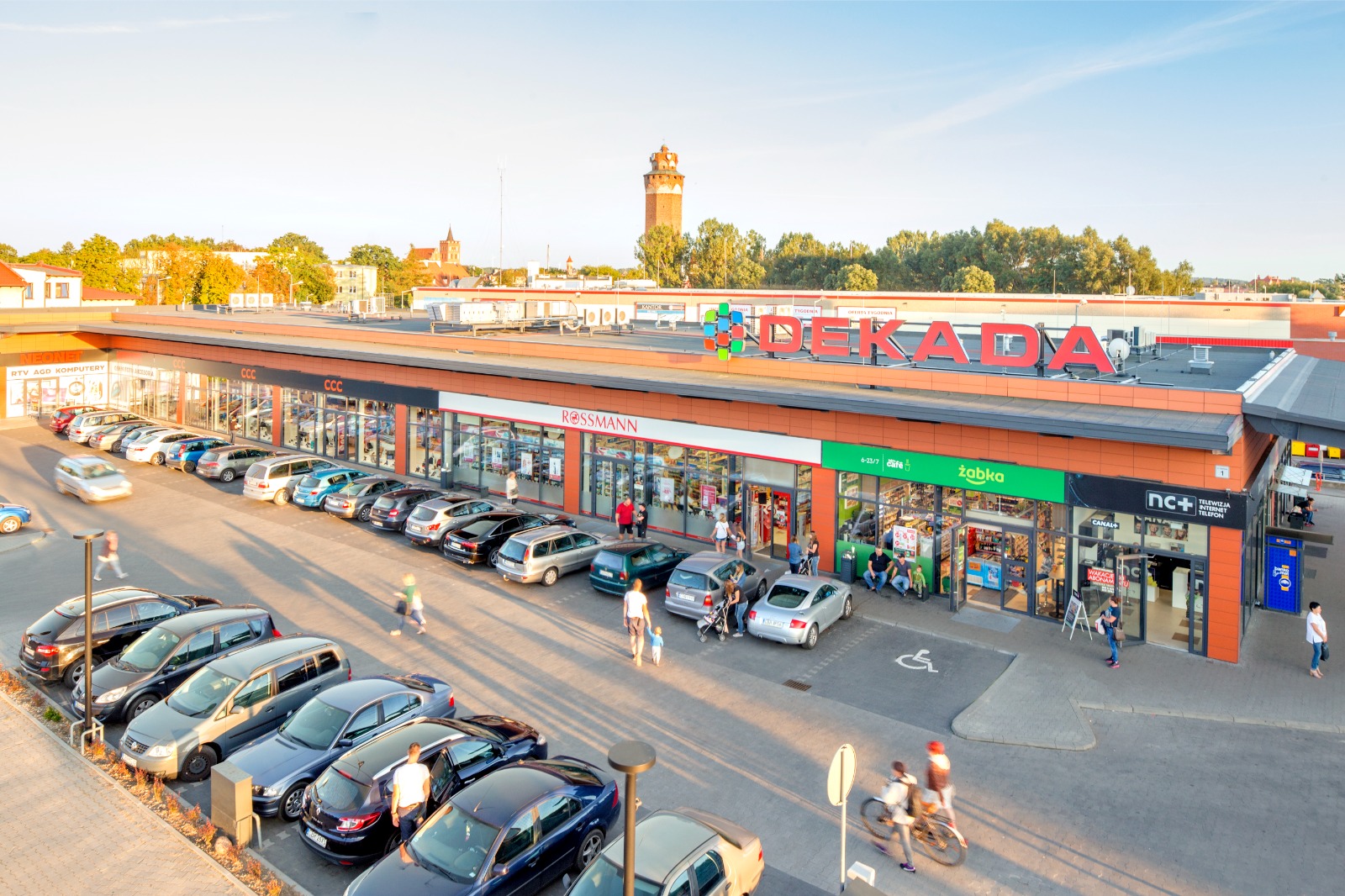 Nowoczesny, parterowy budynek centrum handlowego o pomarańczowej elewacji z napisem „DEKADA” na dachu, parkingiem i witrynami sklepów.