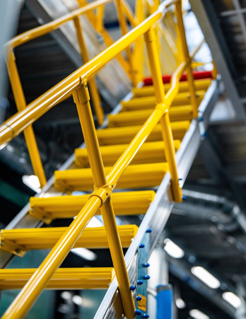 Metalowe schody przemysłowe o intensywnie żółtych stopniach i poręczach prowadzą stromo w górę, w głąb hali. Konstrukcja nośna wykonana jest z matowego aluminium, a całość osadzona w surowym, technicznym wnętrzu z widocznymi instalacjami pod sufitem. Detale obejmują niebieskie śruby mocujące oraz czerwony element ostrzegawczy na górnym podeście.