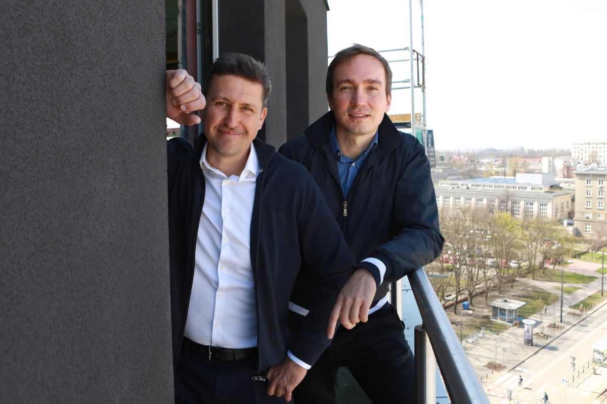Dwaj uśmiechnięci mężczyźni, Piotr Bujnowicz i Szymon Lenkowski, stoją na balkonie nowoczesnego budynku z ciemną elewacją. Jeden w białej koszuli, drugi w niebieskiej, w tle panorama miasta z parkiem i budynkami, widoczne fragmenty rusztowań.