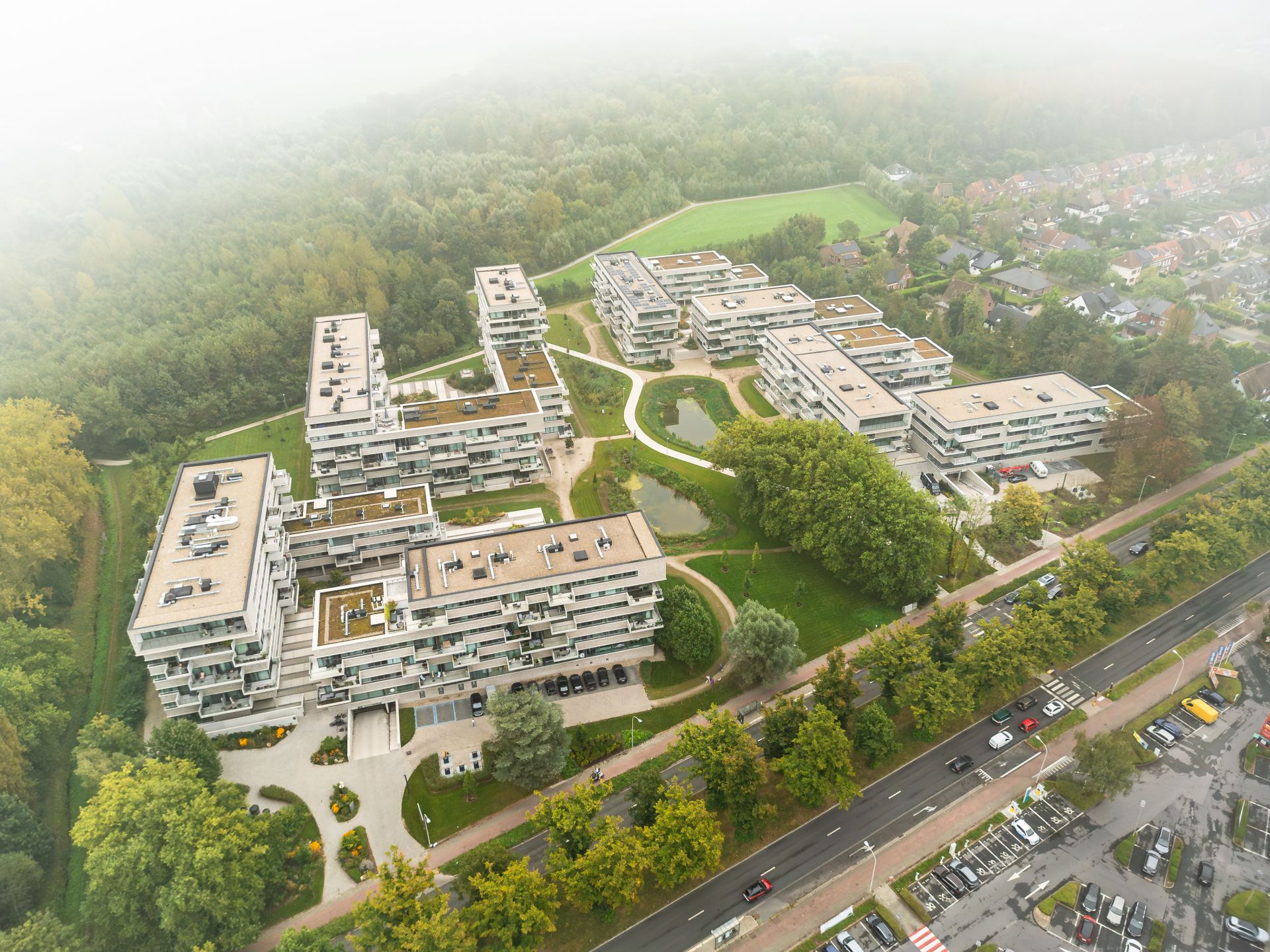 Nowoczesny kompleks mieszkalny o białych elewacjach i kaskadowej architekturze, otoczony lasem i zielenią, z panelami fotowoltaicznymi na dachach.