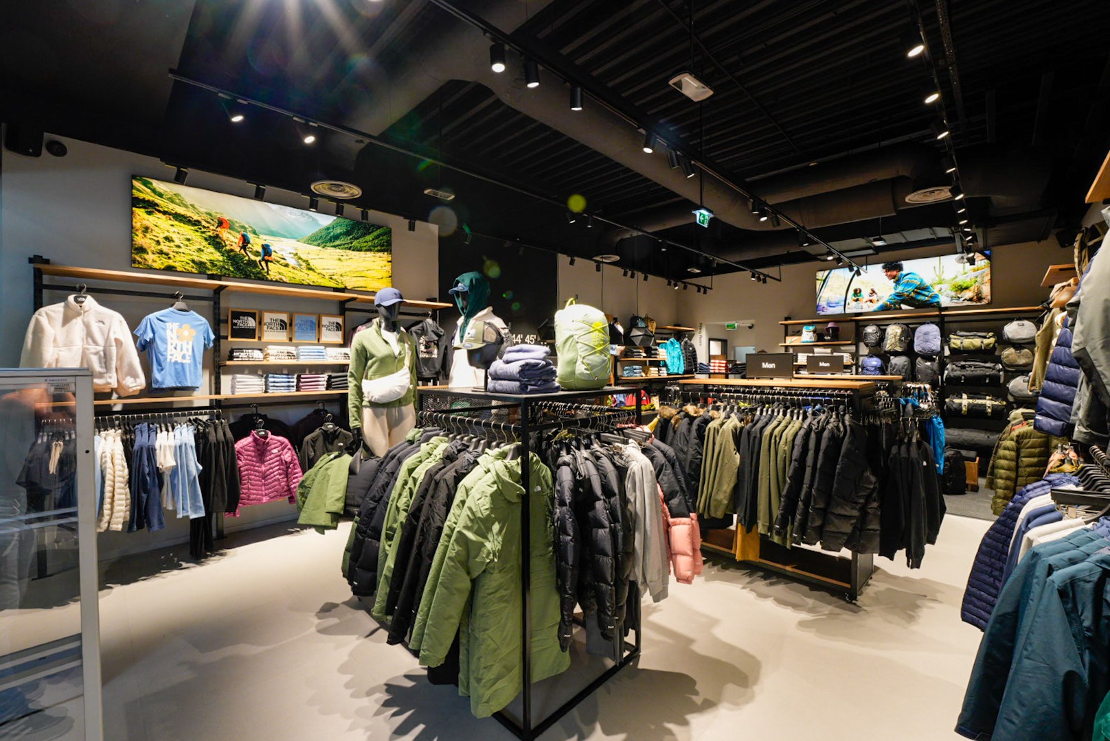 Nowoczesny sklep z odzieżą outdoorową, wieszaki z kurtkami, bluzami i plecakami, manekiny ubrane w sportowe zestawy. Industrialny styl wnętrza.