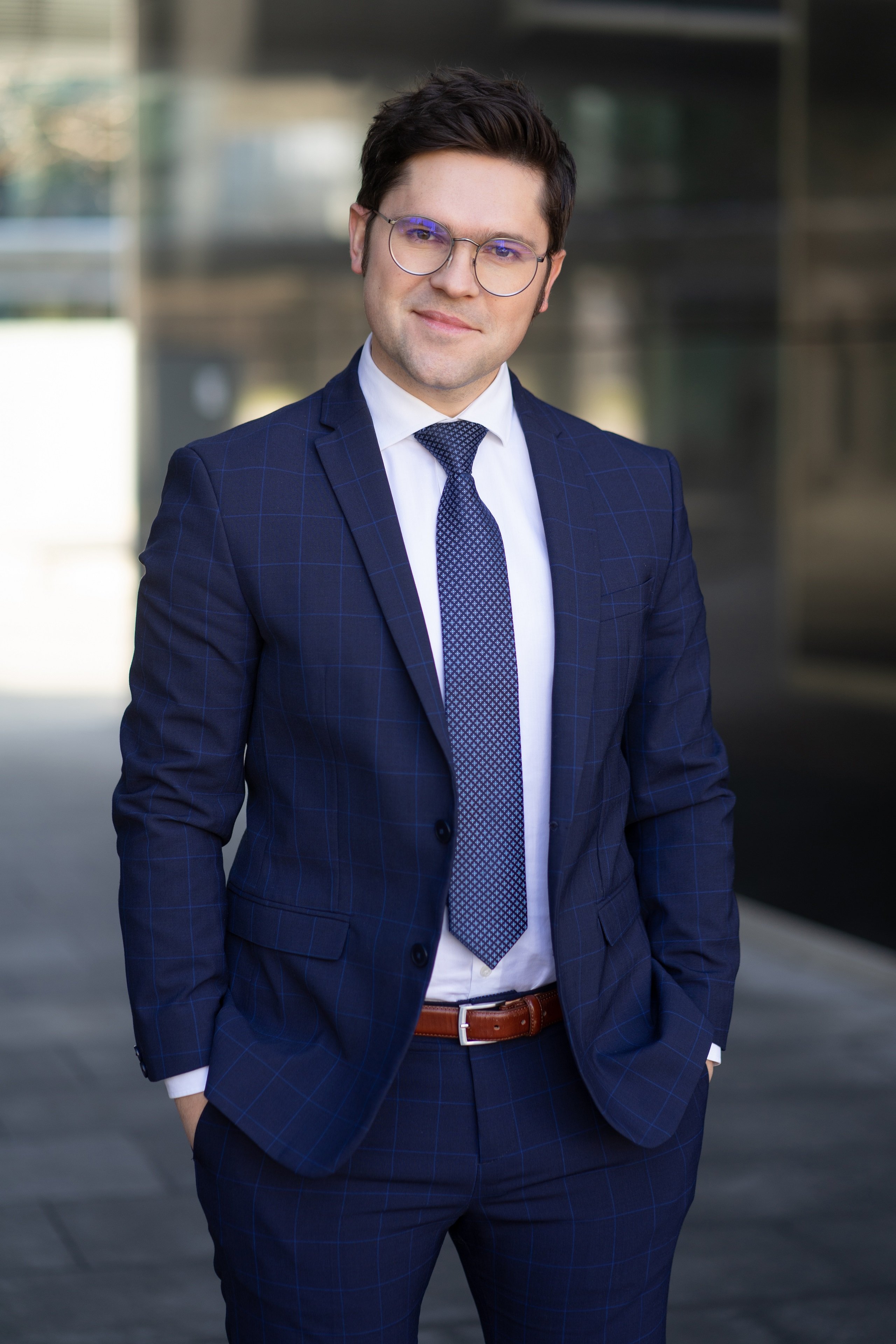 Mężczyzna w okularach korekcyjnych w granatowym garniturze stoi z rękami w kieszeniach na tle biurowca.