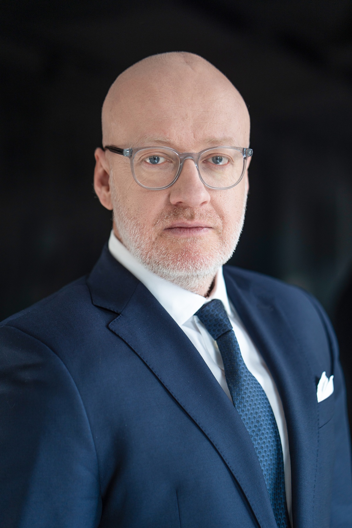 Portret łysego mężczyzny z krótkim siwym zarostem w ciemnoniebieskiej marynarce, białej koszuli, wzorzystym granatowym krawacie i okularach.