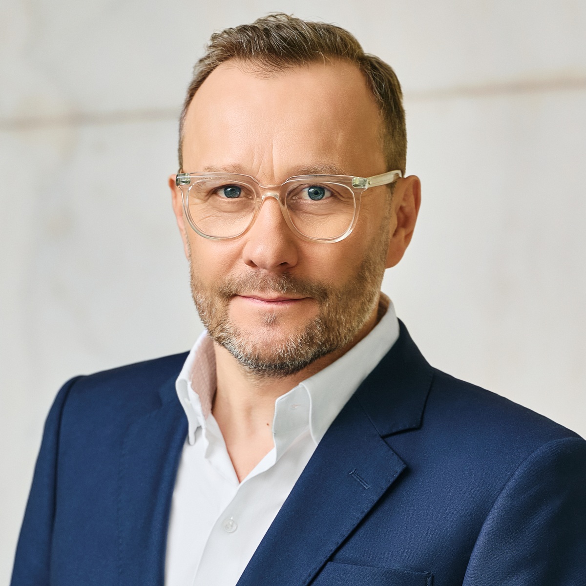 Portret mężczyzny w średnim wieku z krótkimi, ciemnymi włosami, zadbanym zarostem i okularami w przezroczystych oprawkach.