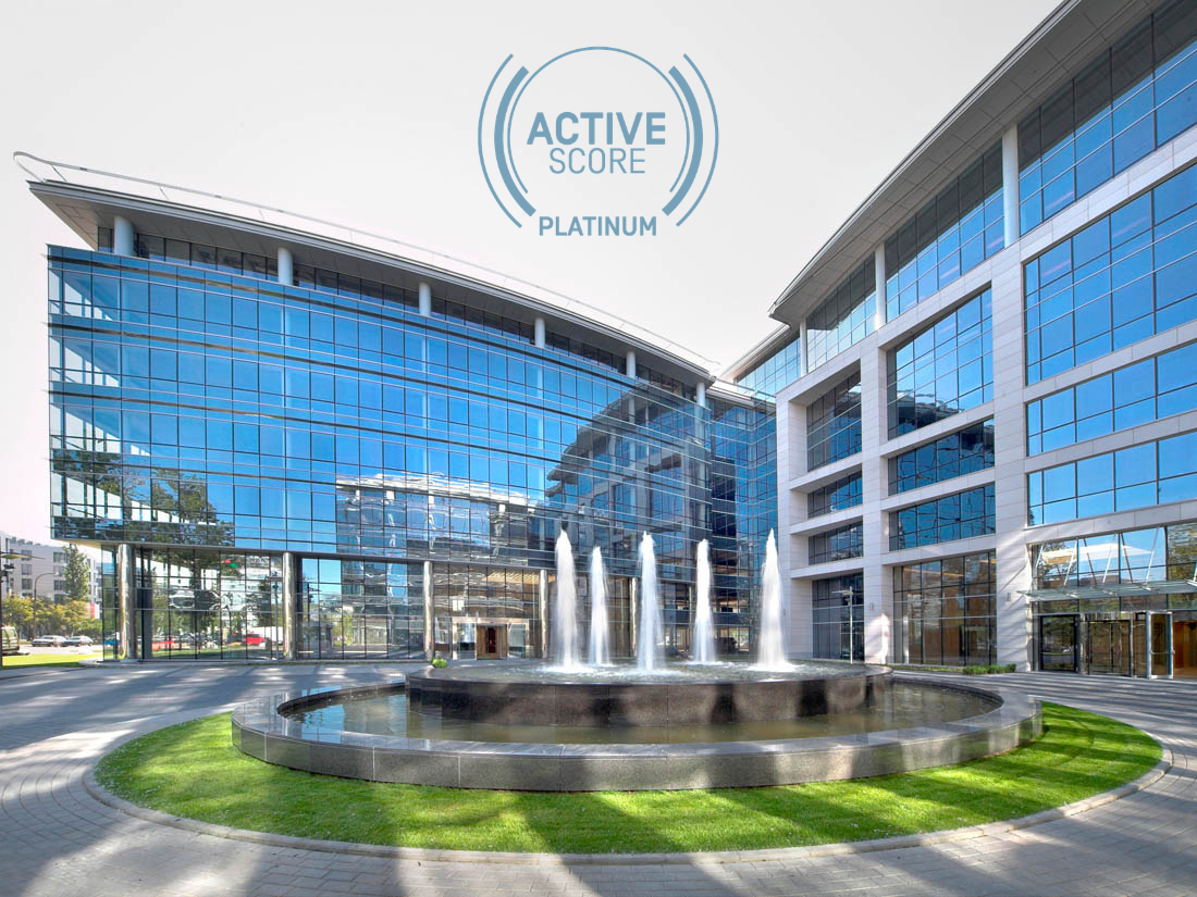 Nowoczesny, przeszklony biurowiec o geometrycznej bryle z okrągłą fontanną i logo ActiveScore Platinum na tle nieba.