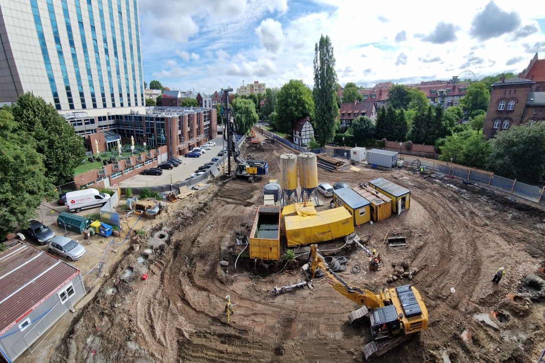 Plac budowy w centrum miasta z rozkopaną ziemią, ciężkim sprzętem, żółtą koparką gąsienicową oraz wysoką palownicą.