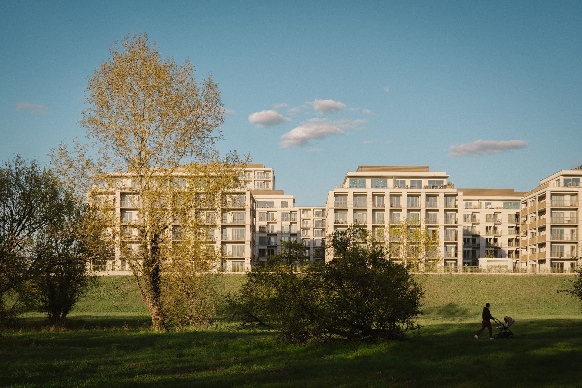 Wielokondygnacyjny kompleks apartamentowców o jasnych elewacjach, dominujący nad zielonym terenem rekreacyjnym.