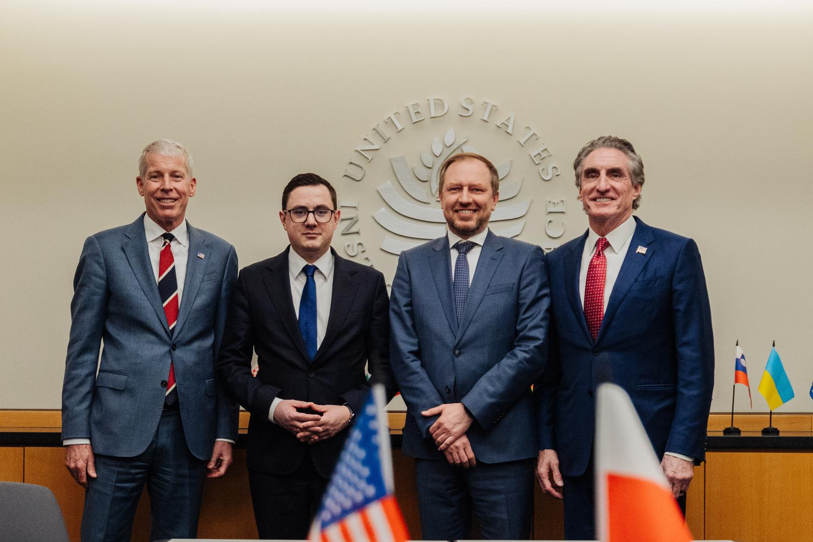 Czterech mężczyzn w garniturach stoi w rzędzie na tle jasnej ściany z godłem United States Institute of Peace.