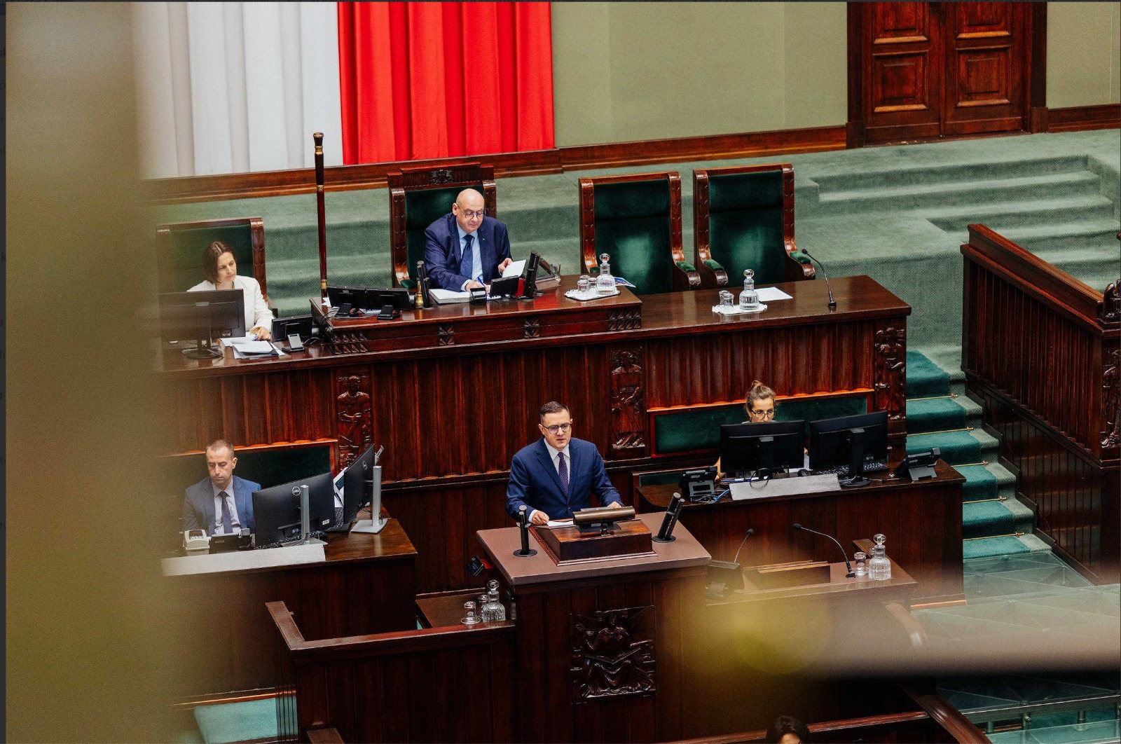 Wnętrze sali plenarnej parlamentu z mężczyzną w granatowym garniturze przemawiającym z mównicy.