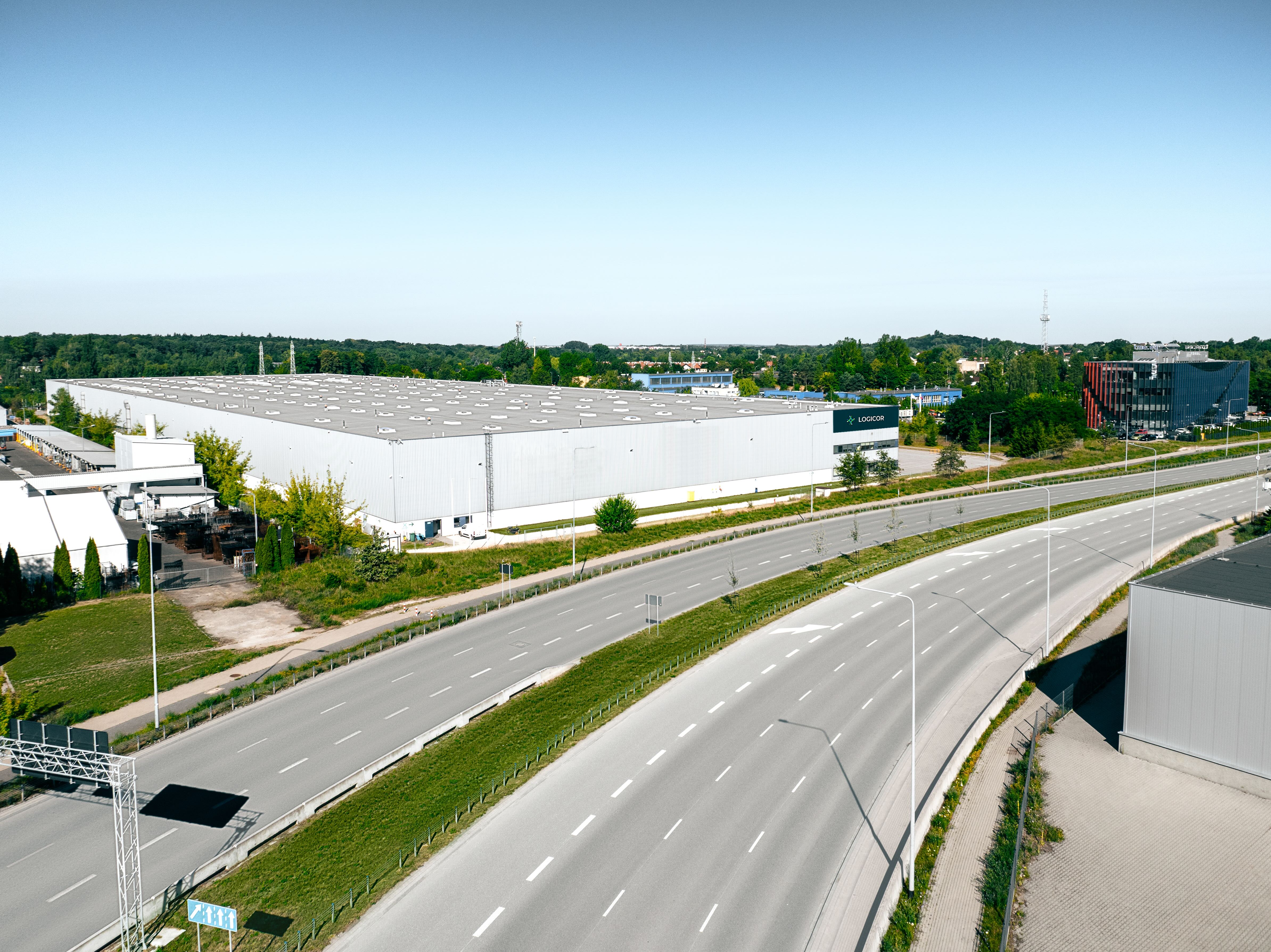 Rozległa, jasnoszara hala magazynowa Logicor z napisem „LOGICOR” obok wielopasmowej arterii drogowej i nowoczesnego budynku biurowego.