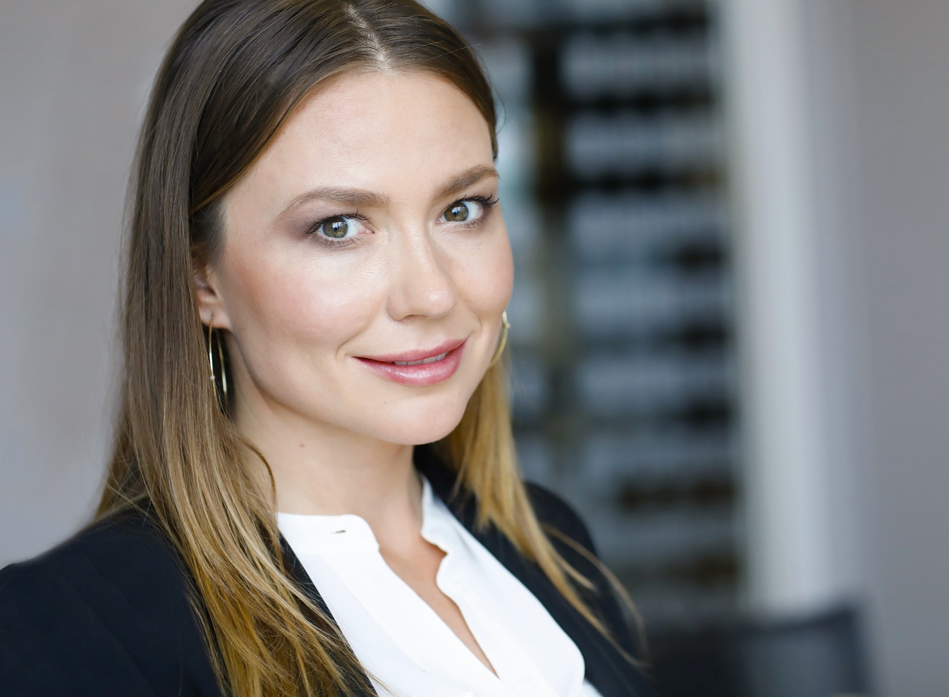 Portret młodej kobiety o brązowych włosach, jasnych oczach i złotych kolczykach w kształcie kół. Uśmiecha się, ubrana w białą bluzkę i czarną marynarkę.