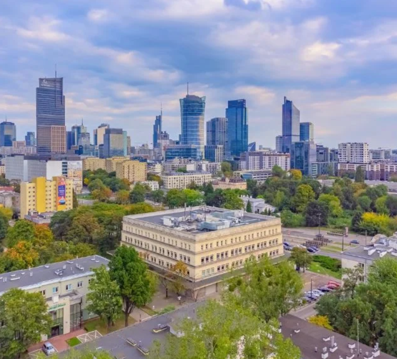 Panorama miasta z niską zabudową i nowoczesnym centrum biznesowym. Na pierwszym planie jasnopiaskowy budynek otoczony zielenią, w tle wieżowce.