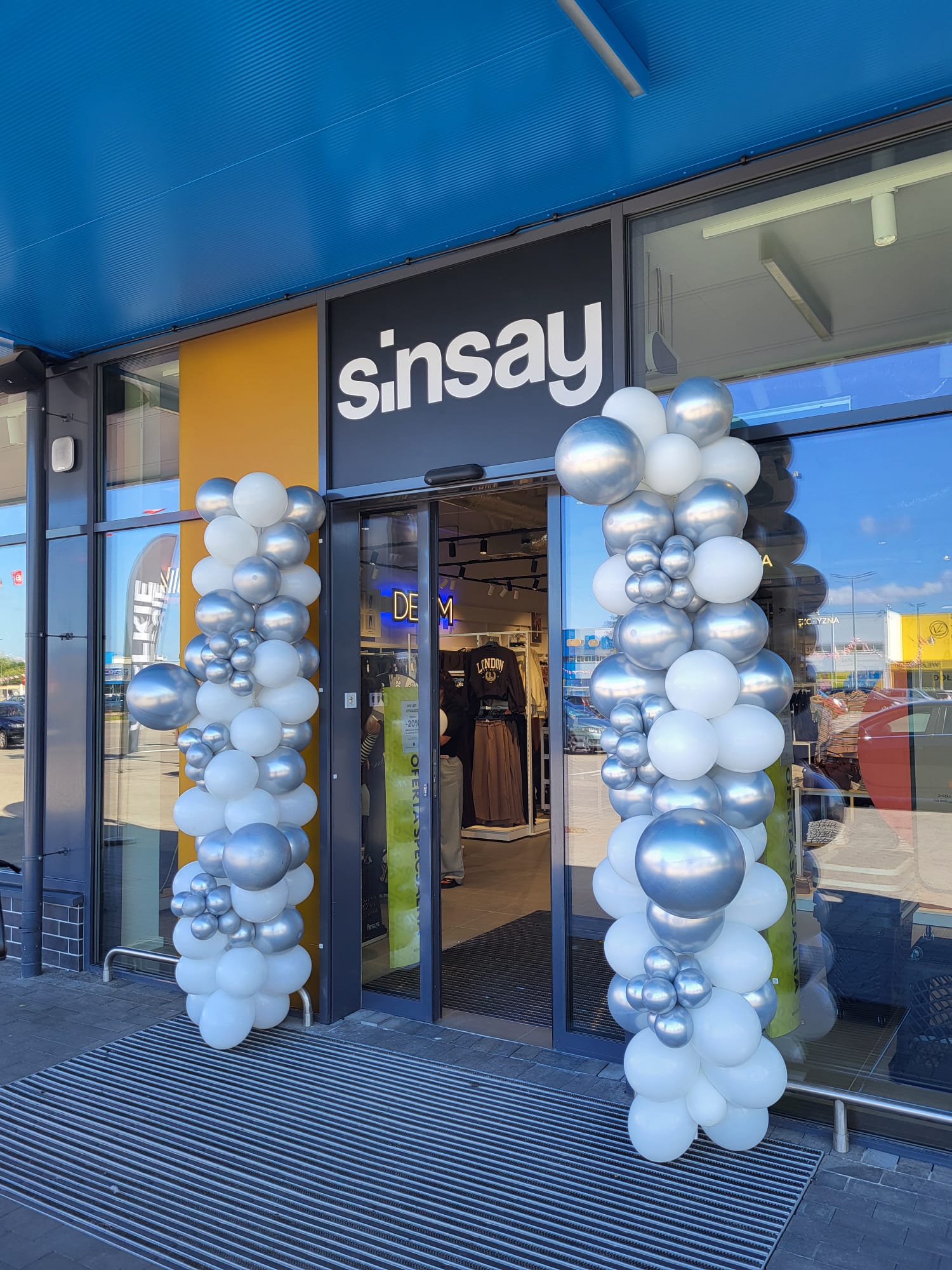 Wejście do sklepu Sinsay z pionowymi girlandami z białych i srebrnych balonów, widoczne regały z ubraniami i neonowy napis DENIM w środku.