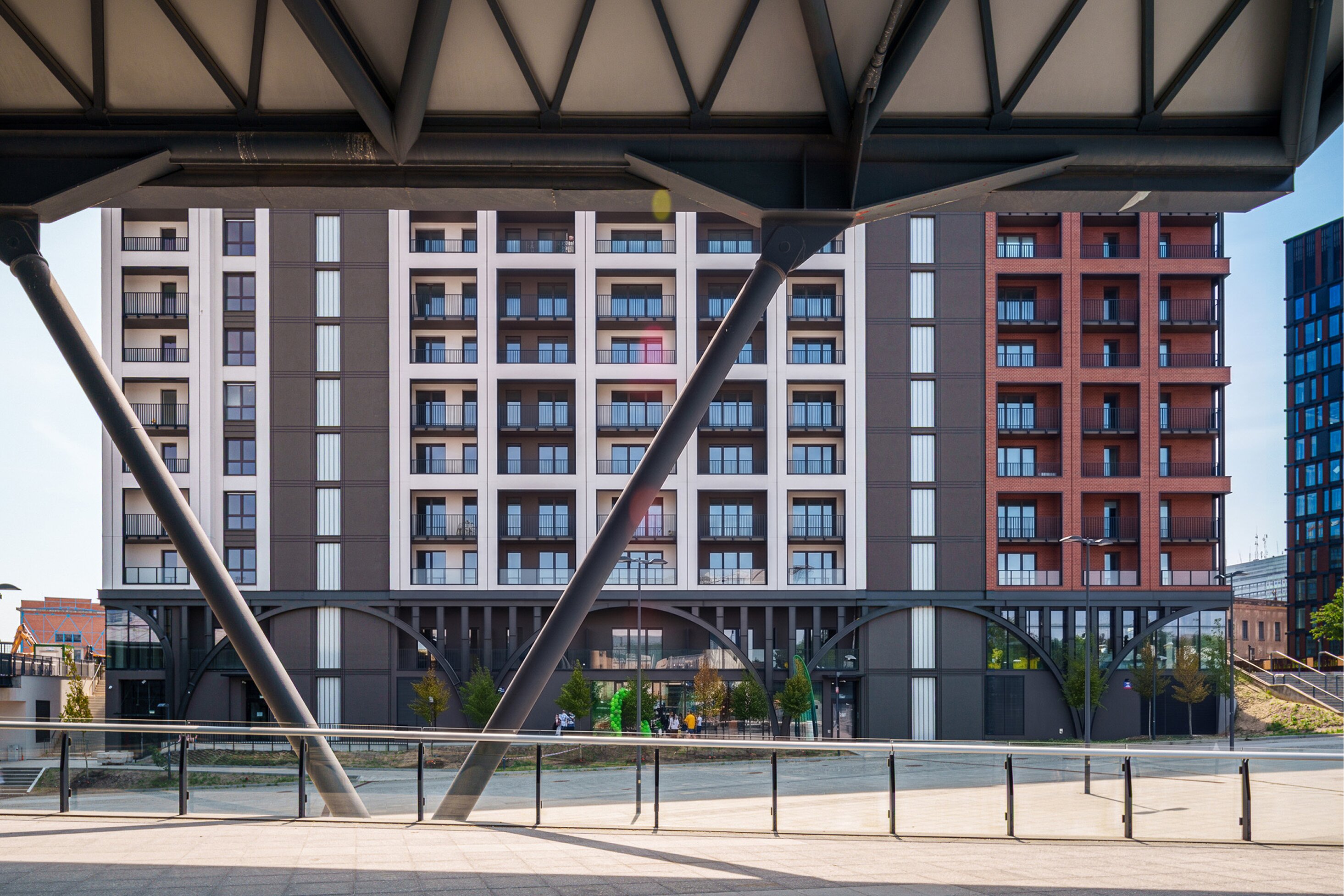 Wielokondygnacyjny budynek o nowoczesnej architekturze z białymi, grafitowymi i ceglanymi elewacjami. Nad nim stalowe zadaszenie.