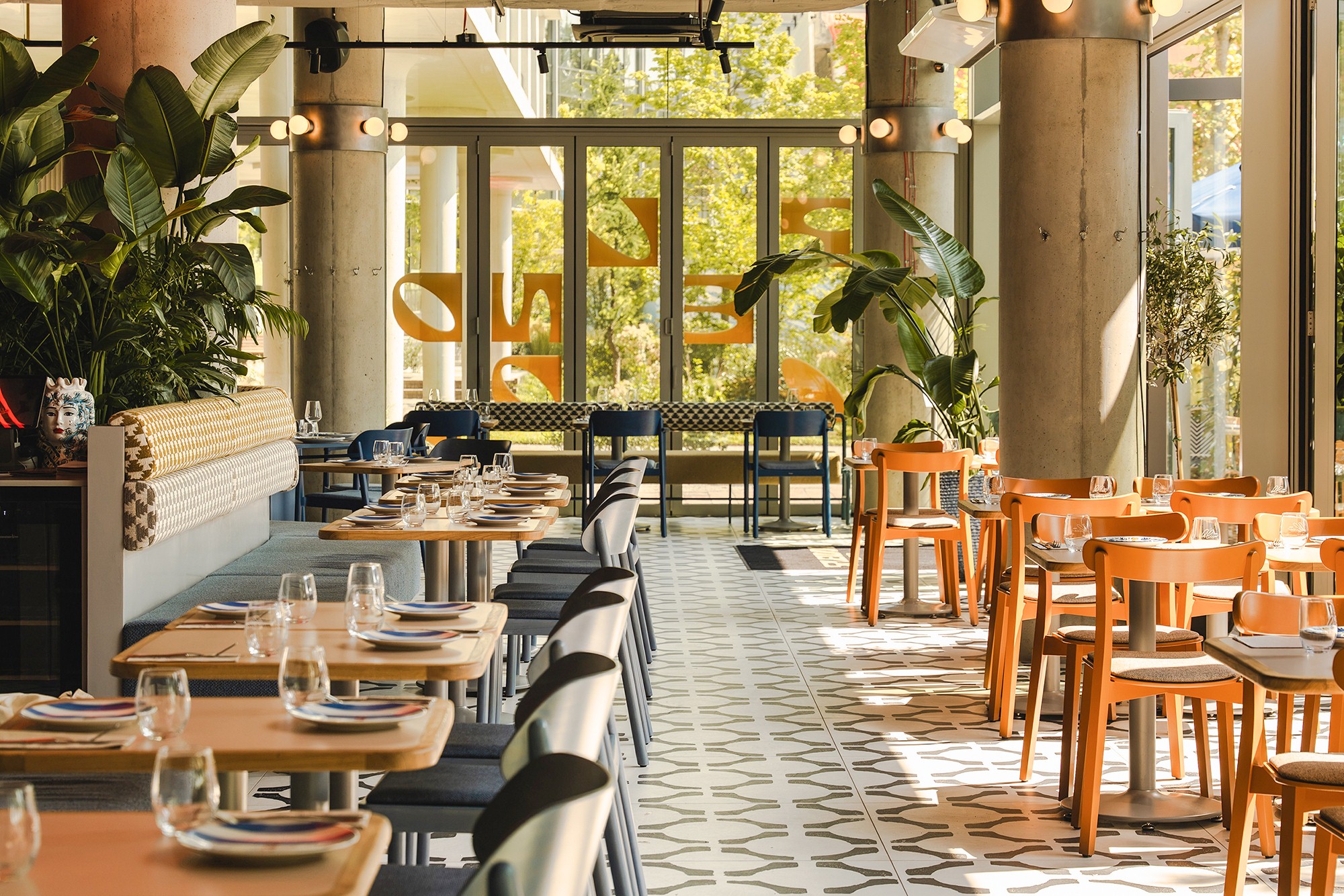 Nowocześnie urządzone wnętrze restauracji z betonowymi słupami, czarno-białą podłogą, drewnianymi stolikami i kolorowymi krzesłami.