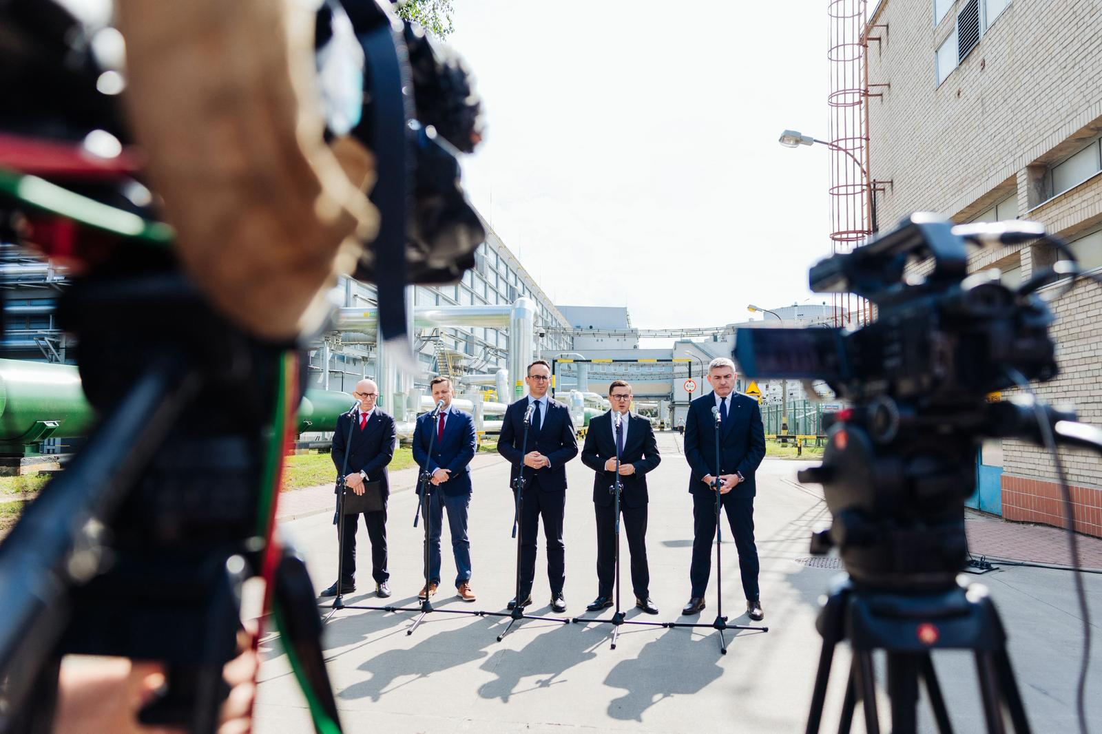 Pięciu mężczyzn w garniturach stoi przed mikrofonami na tle infrastruktury przemysłowej, filmowanych przez kamery.