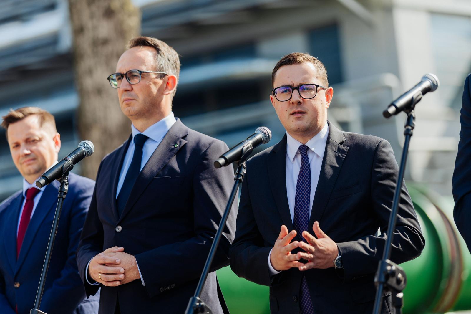 Trzech mężczyzn w garniturach stoi przed mikrofonami, jeden z nich, w okularach, przemawia z ożywioną gestykulacją.