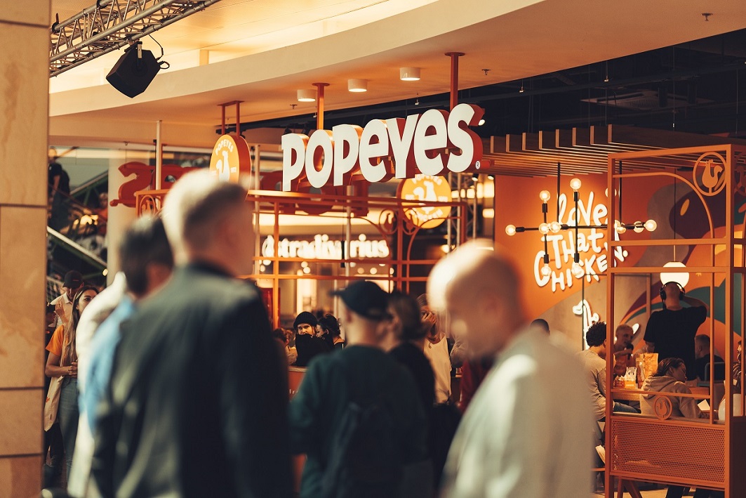 Wejście do restauracji Popeyes w centrum handlowym z widocznym dużym logotypem na pomarańczowym tle i oświetlonym wnętrzem.